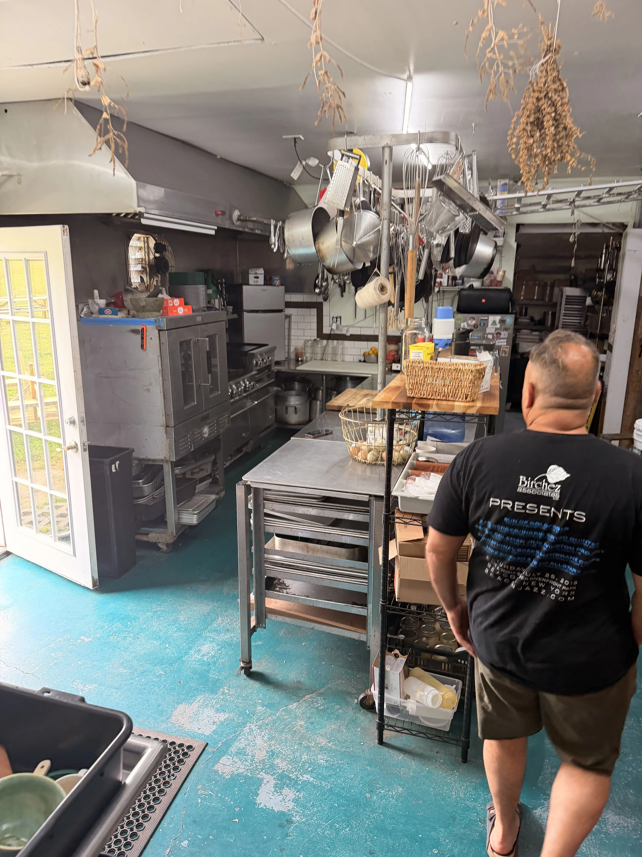 A kitchen with hanging herbs and utensils, a man with short hair and a black T-shirt walking in the foreground, and various kitchen appliances and storage shelves in the background.