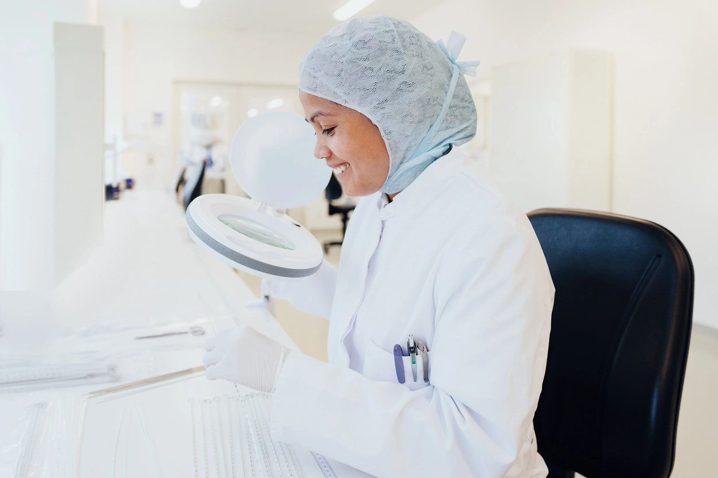 The image shows a person wearing protective laboratory clothing, including a white lab coat, disposable gloves, and a light blue hair cover. The individual is seated at a workstation in a clean, bright laboratory environment.