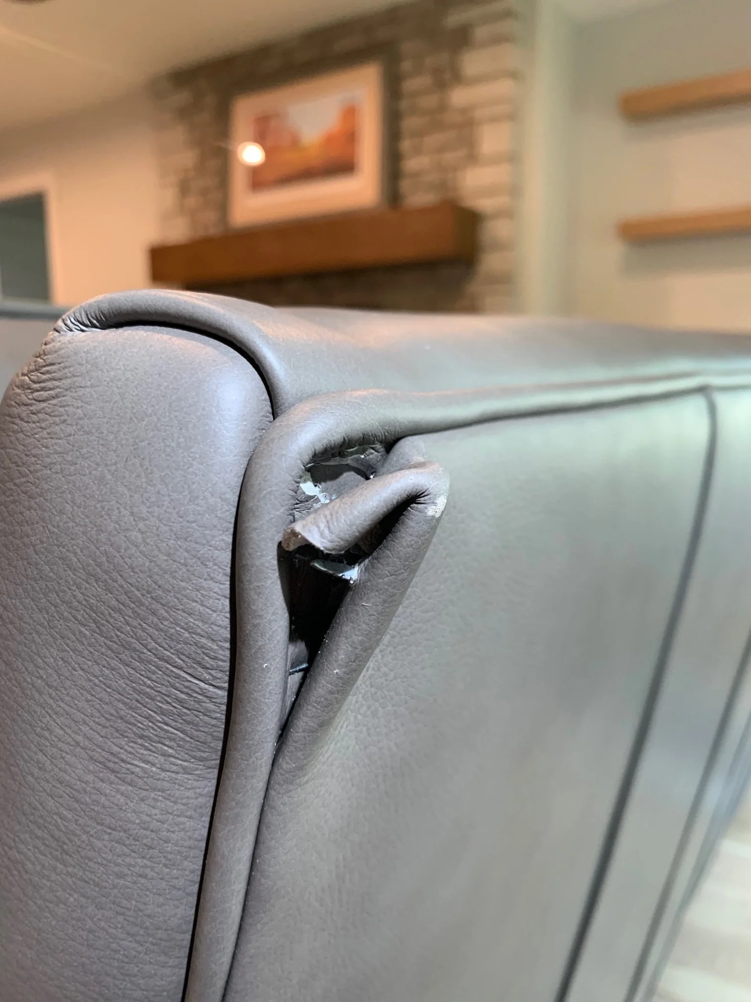 Close-up of a beige upholstered armrest of a chair with a damaged area showing padding and exposed metal parts, set against a background of a living room with brick fireplace and framed picture.