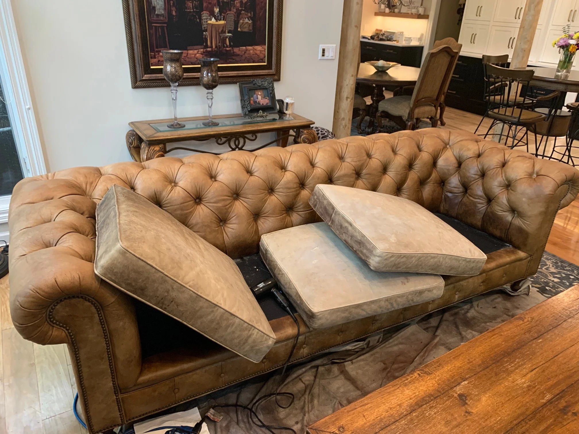Living room with a large tan leather tufted sofa, a wooden side table with candles and a picture frame, a dining area with a round table and chairs, and a kitchen with white cabinets and a vase of flowers.