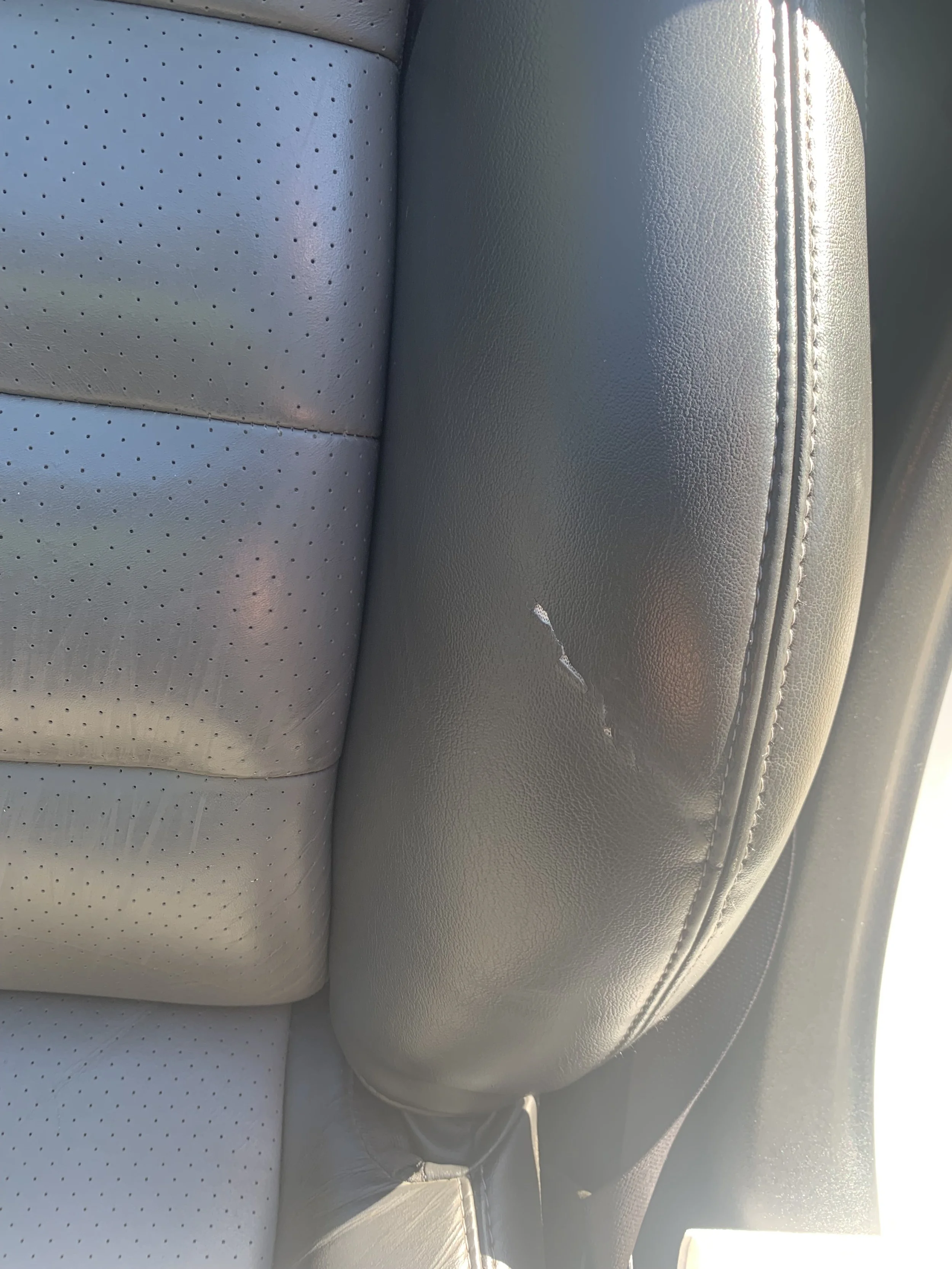 A car interior showing a light-colored perforated leather seat with visible stitching and a small white speck of damage or dirt.
