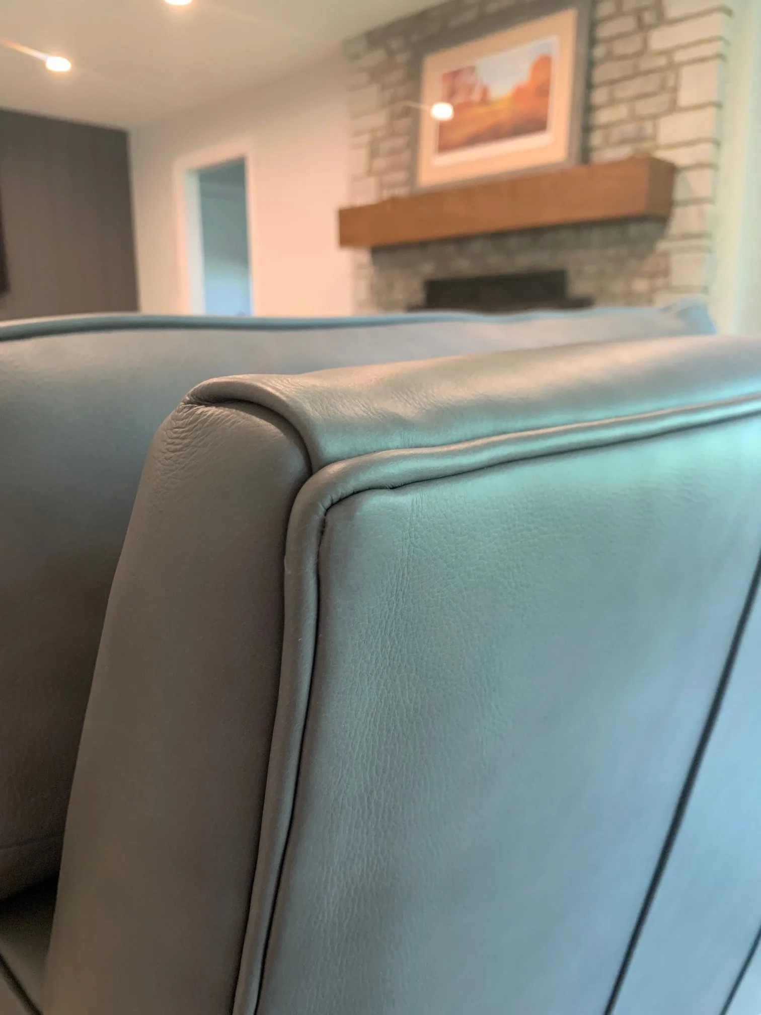 Close-up of a beige leather armrest of a sofa with a background of a living room wall, fireplace, brick wall, and framed painting.