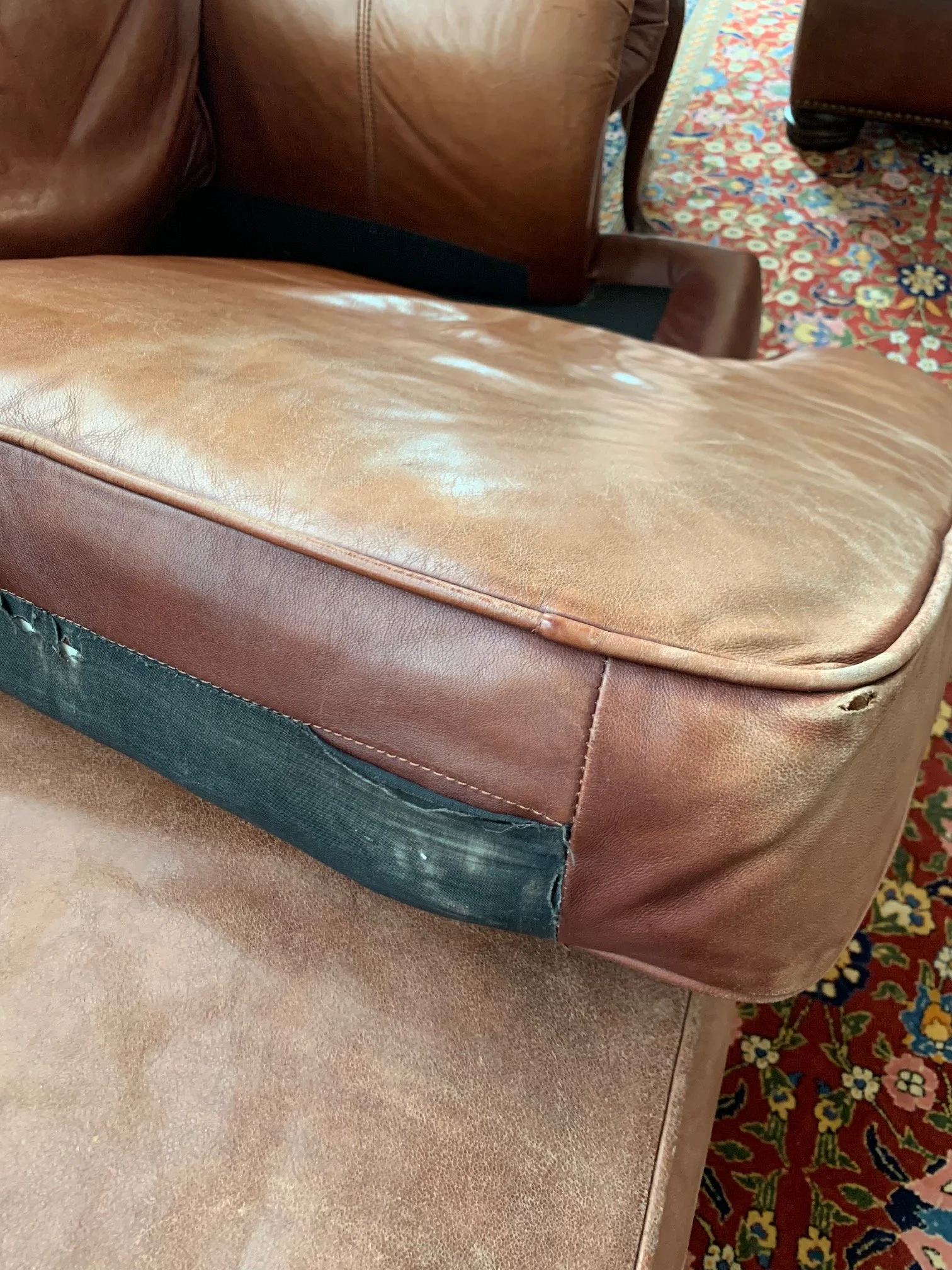 Close-up of a worn leather armrest on a brown chair, with visible scuffs and tears, next to a reddish patterned carpet.