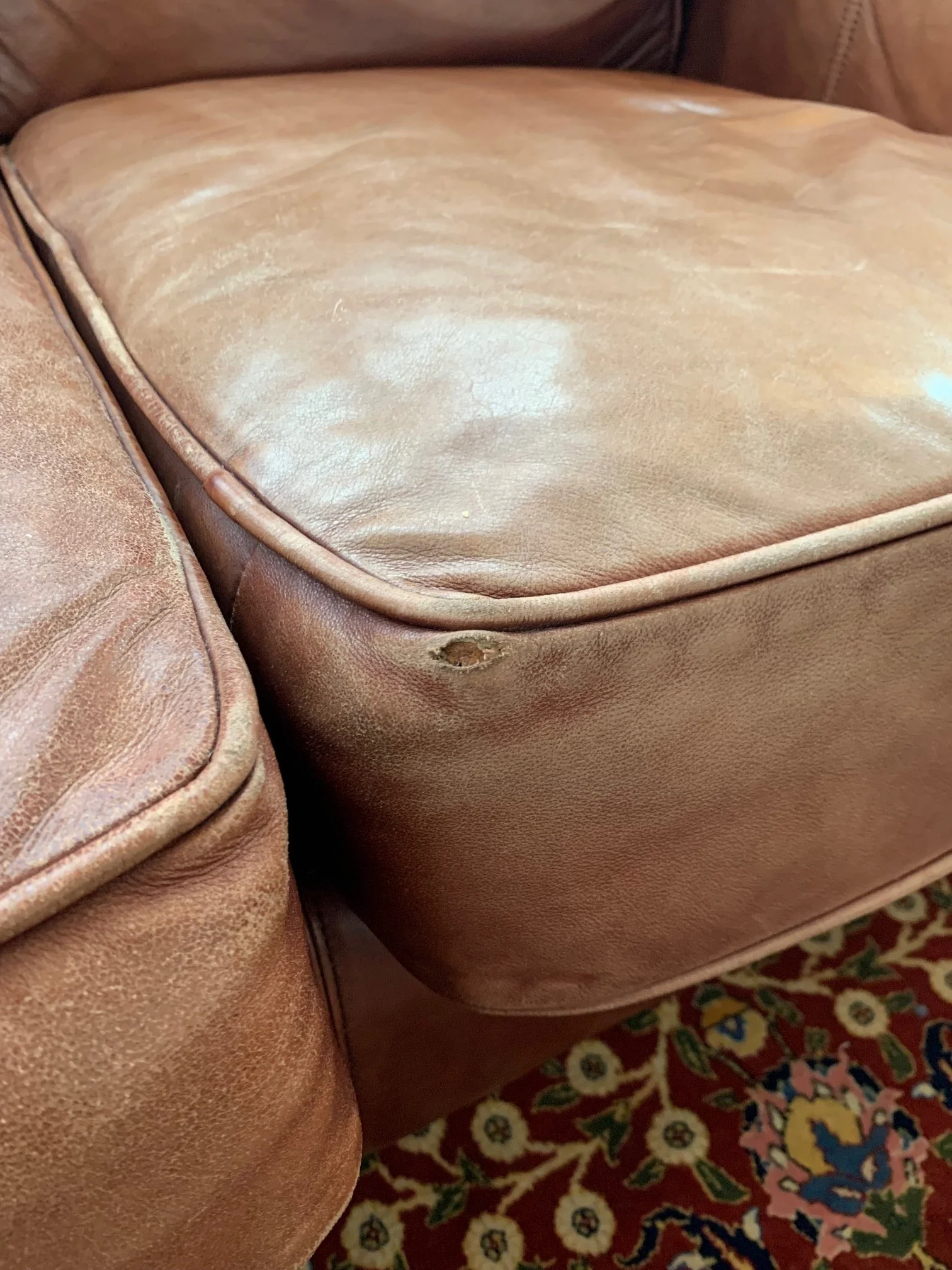 Close-up of a worn, light brown leather sofa armrest with visible scuffs and discoloration, next to a patterned rug with floral and geometric designs.