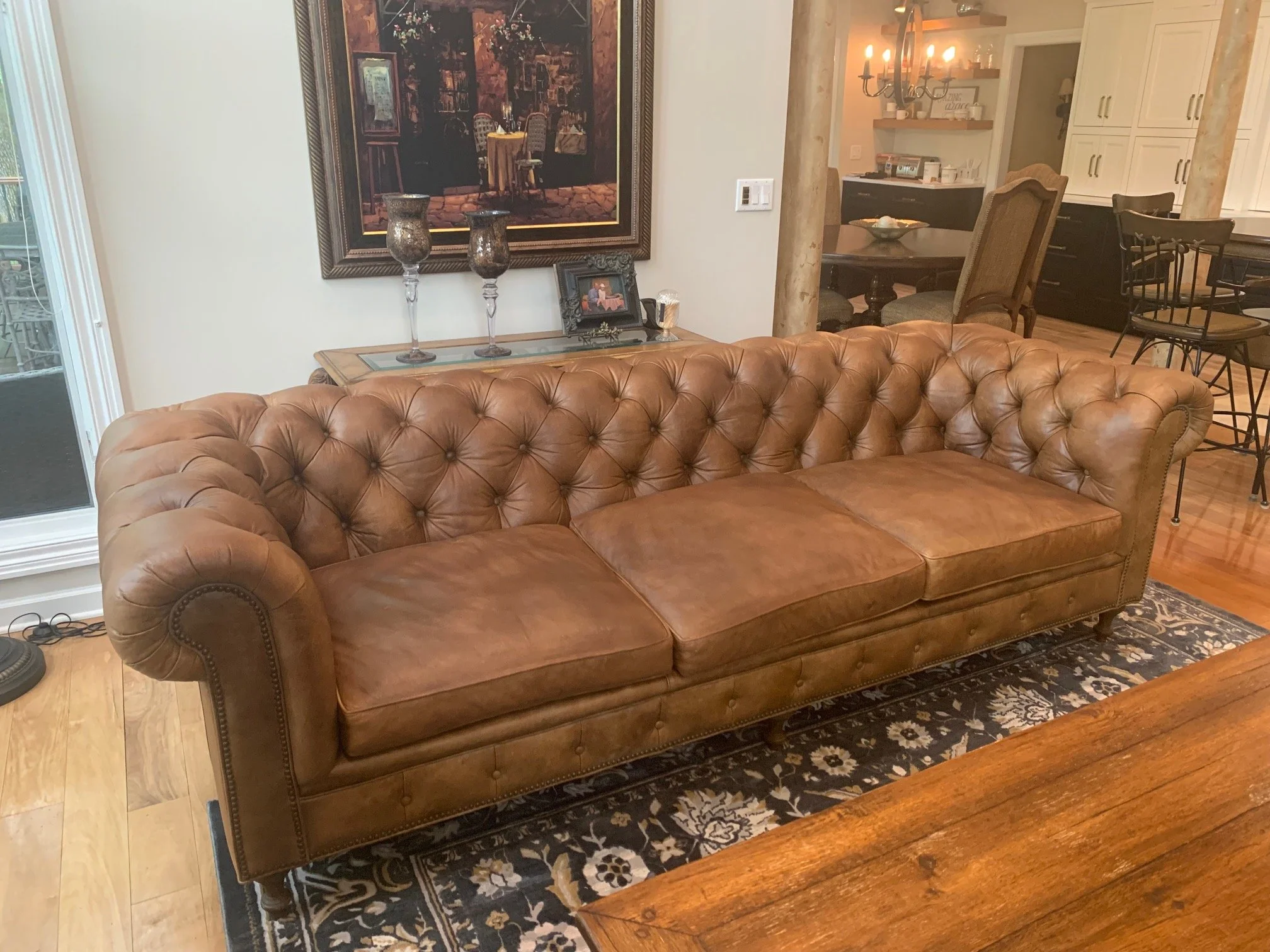 A living room with a brown leather Chesterfield sofa, a wooden coffee table, a black and beige area rug, and a corner with a dining table and chairs. There's a framed painting with flowers hanging on the wall, a console table with vases, a picture frame, and candles. The background shows an open kitchen with white cabinets, a chandelier, countertops, and kitchen appliances. There is also a sliding glass door leading outside.