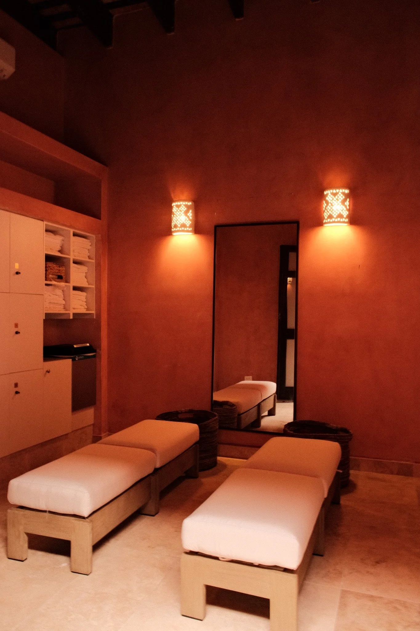 Spa or relaxation room with white lockers, a decorative mirror with an ornate frame, two wall-mounted lamps, and white cushioned seating on wooden stands. The walls are painted a warm terracotta color, and green plants top the lockers.