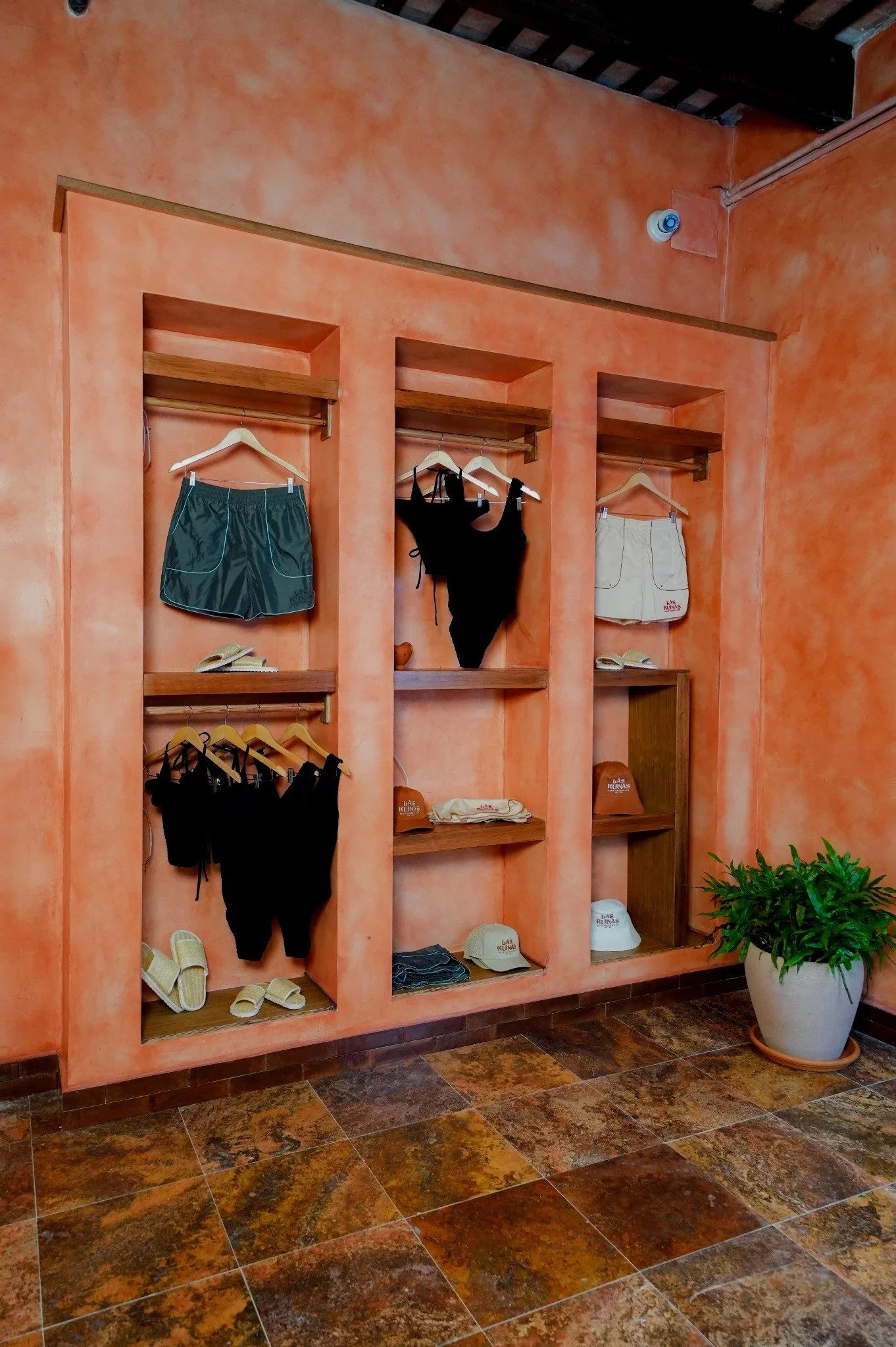 Display of swimwear, hats, and sandals in a coral-colored store with a potted plant on a tiled floor.