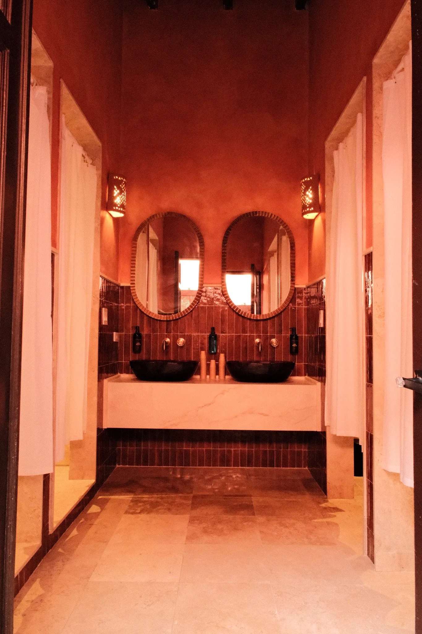 Spa or relaxation room with white lockers, a decorative mirror with an ornate frame, two wall-mounted lamps, and white cushioned seating on wooden stands. The walls are painted a warm terracotta color, and green plants top the lockers.