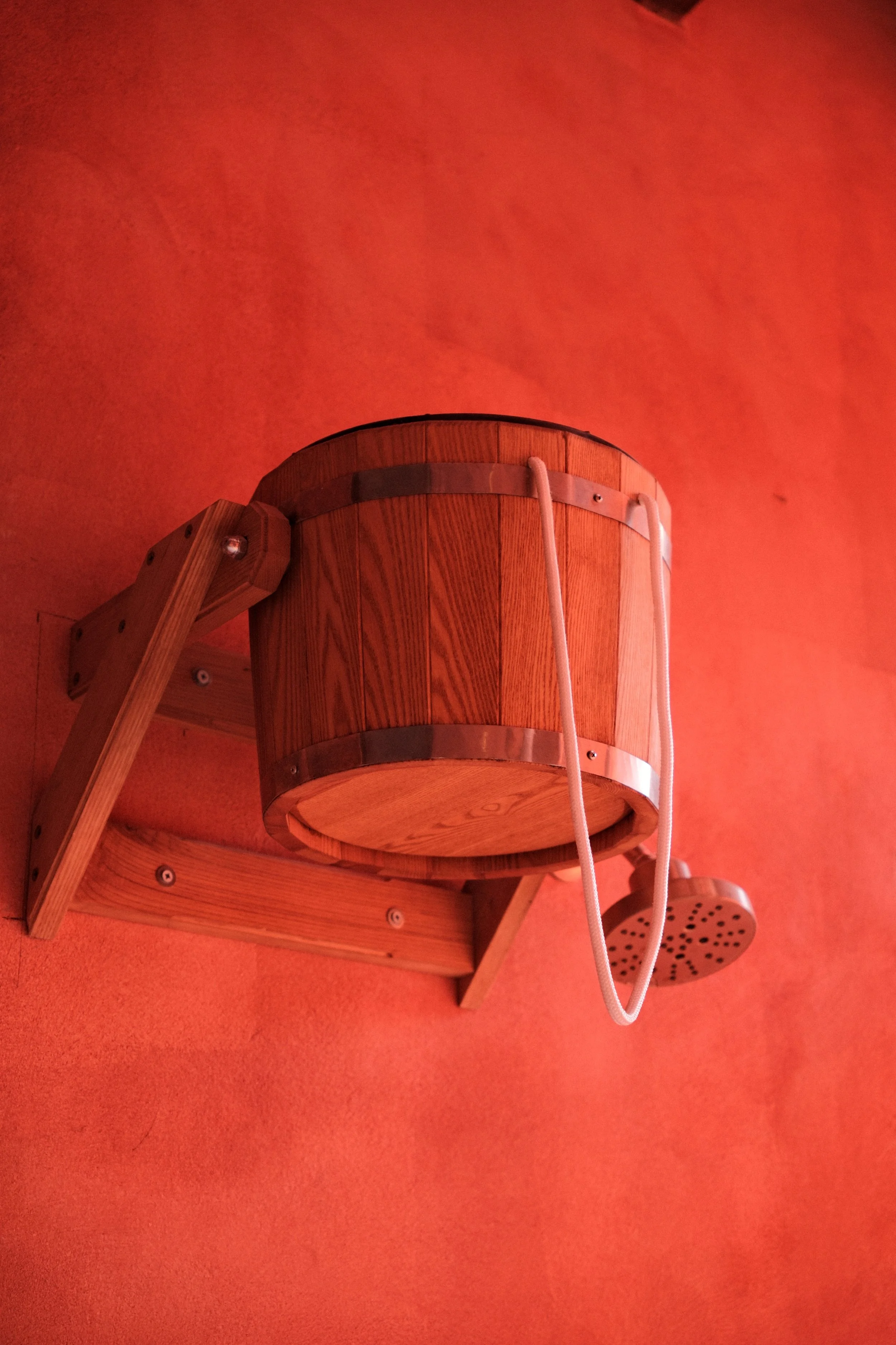 Wooden sauna bucket with a lid, attached to a wall-mounted wooden bracket and a small wooden shower head, set against a red-orange wall.