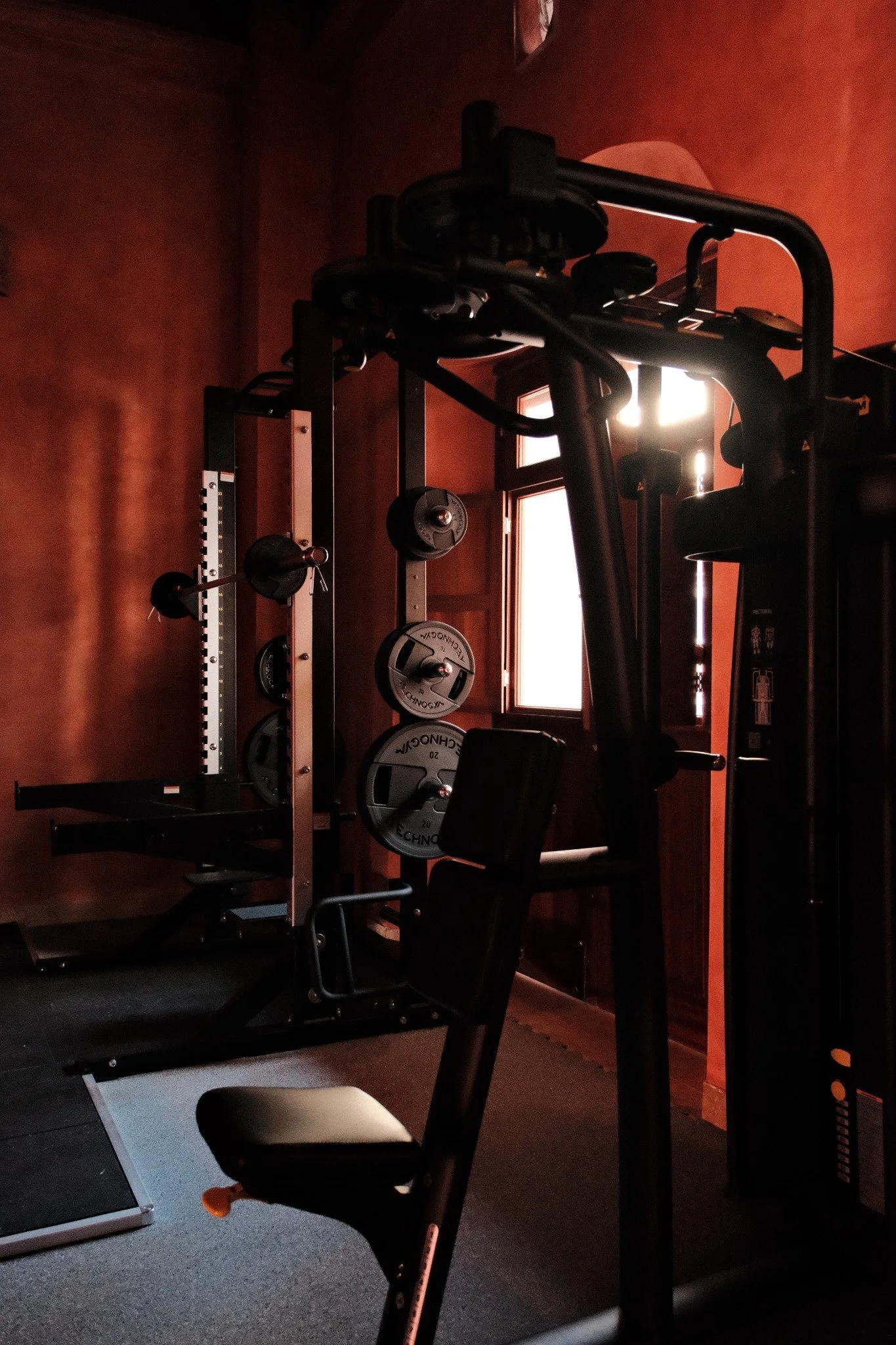 A modern gym with various exercise equipment including treadmills, elliptical machines, and weight lifting stations, with orange textured walls, black ceiling beams, mirrors, and windows with shutters.
