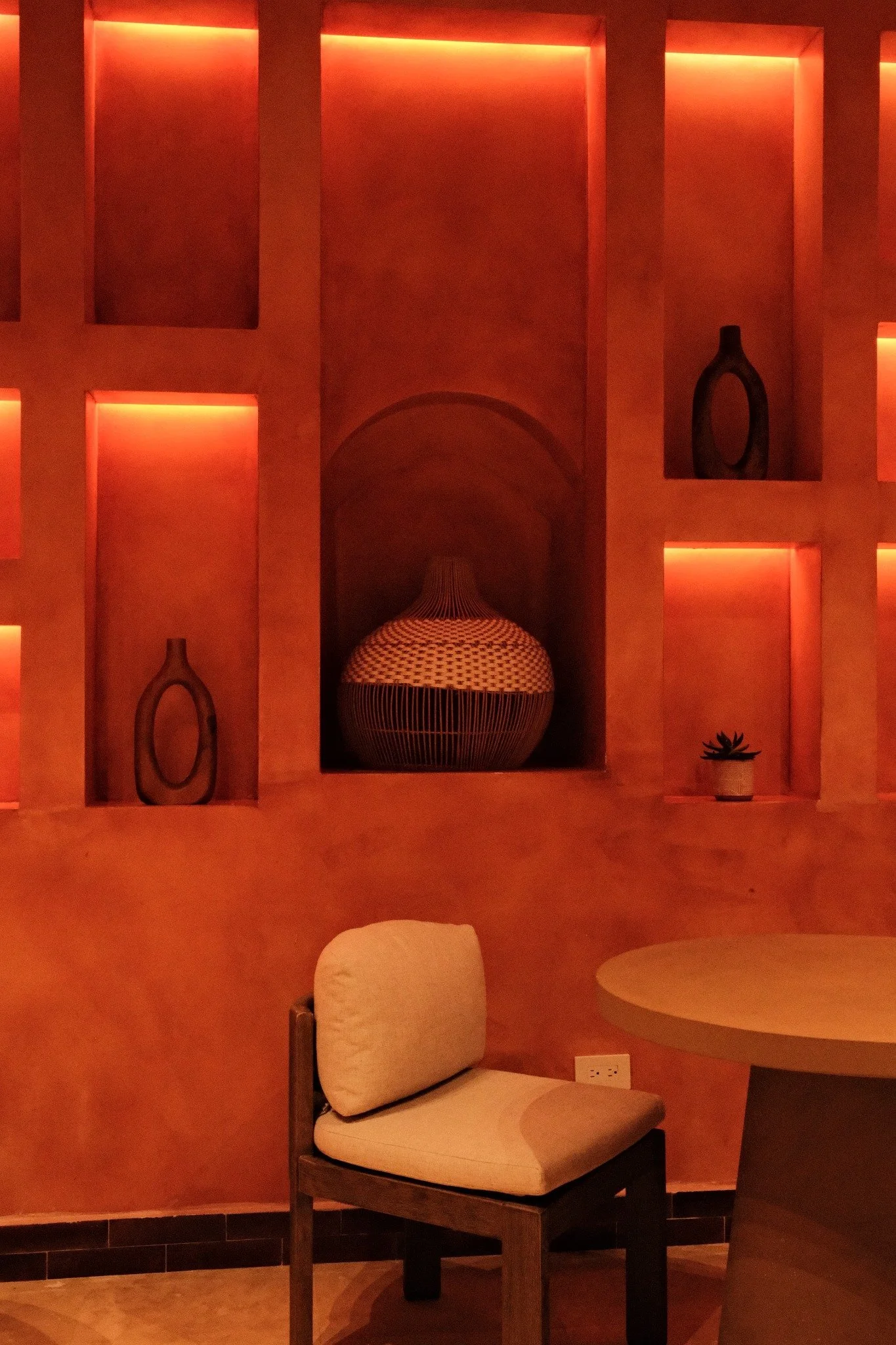 A cozy indoor space with warm orange lighting, built-in wall niches containing vases and a small plant, a beige cushioned chair, and a circular table.