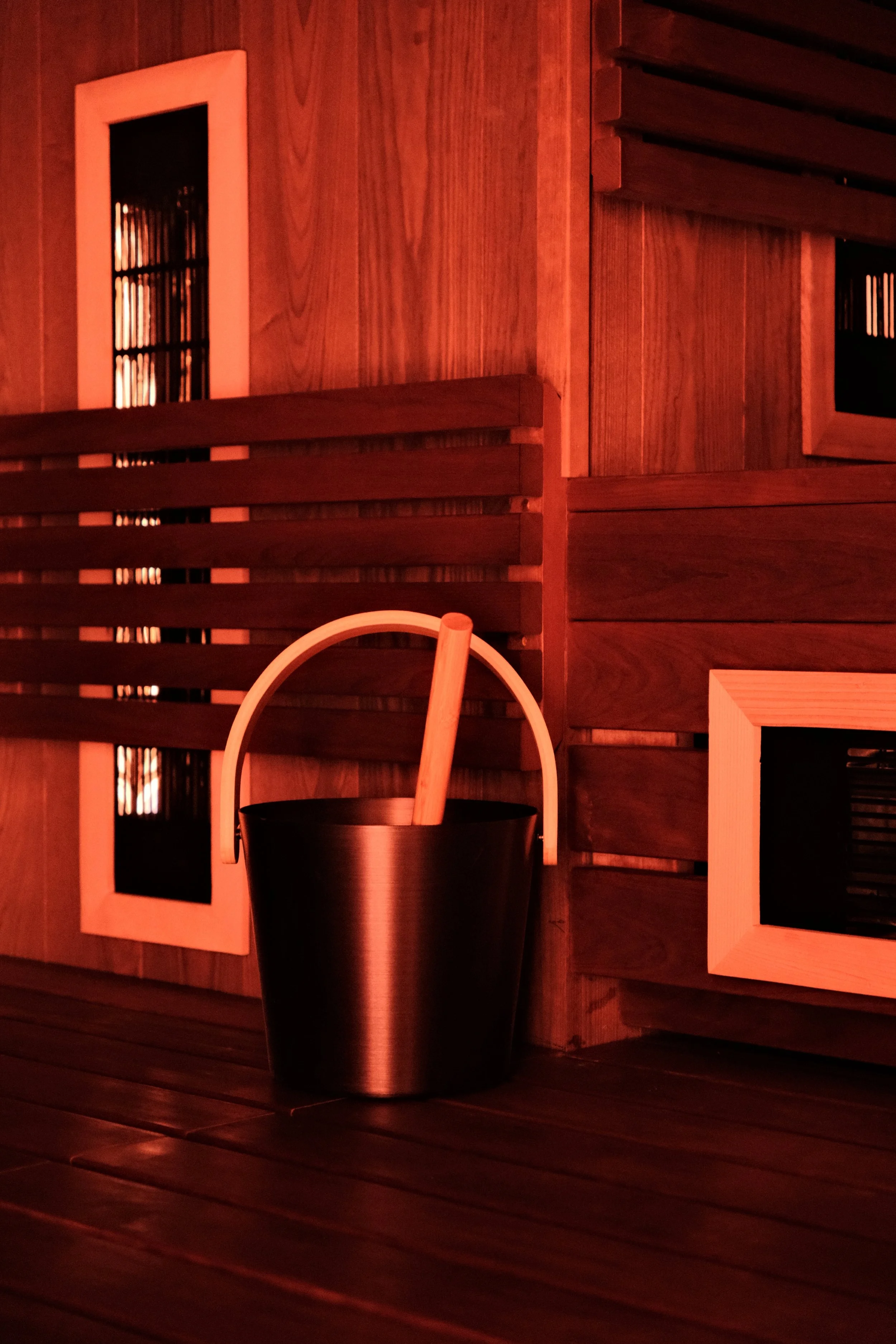 A sauna room with wooden walls and flooring. A black bucket with a wooden ladle inside is placed on the floor. Wooden slats are arranged across the walls, with some framed panels showing darkened spaces, likely for heating or ventilation.