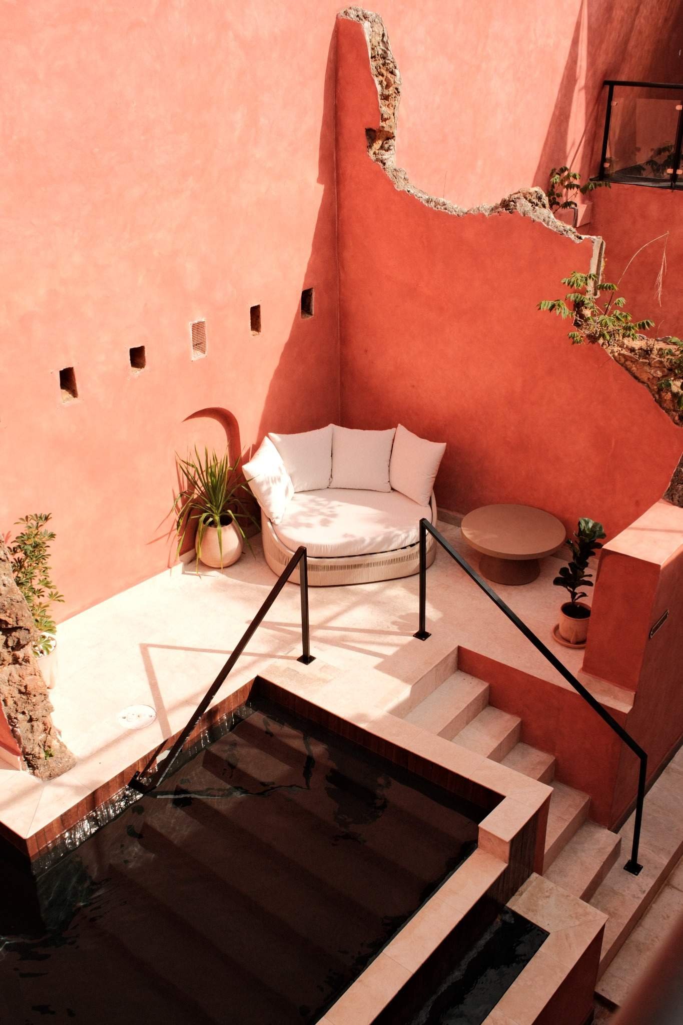 Interior space with a pink wall, a small round white sofa with pillows, potted plants, and stairs adjacent to a black railing.