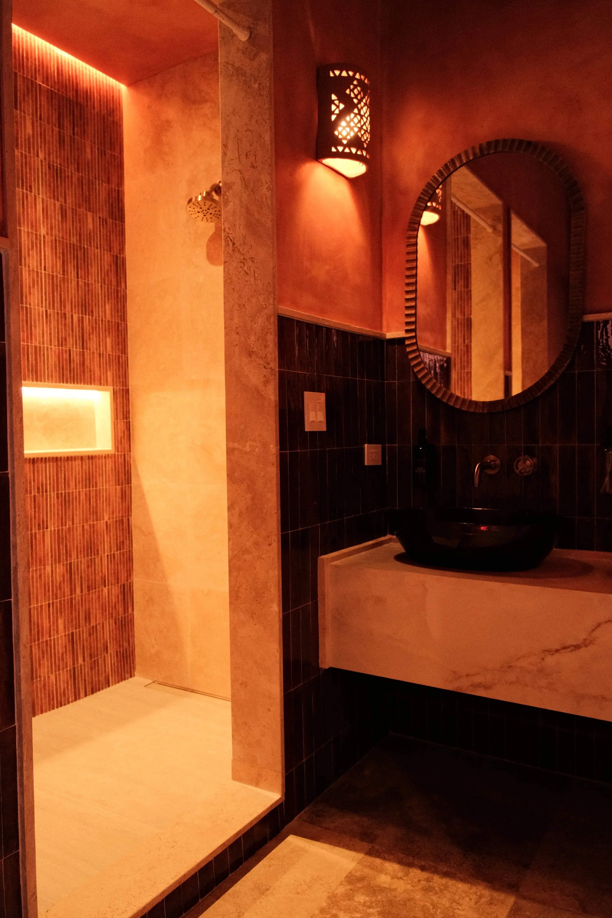 A dimly lit bathroom with a shower area on the left with textured tile walls, an inset shelf, and a rain shower head. On the right, there is a black vessel sink with a marble counter, an oval mirror, and wall-mounted lights.