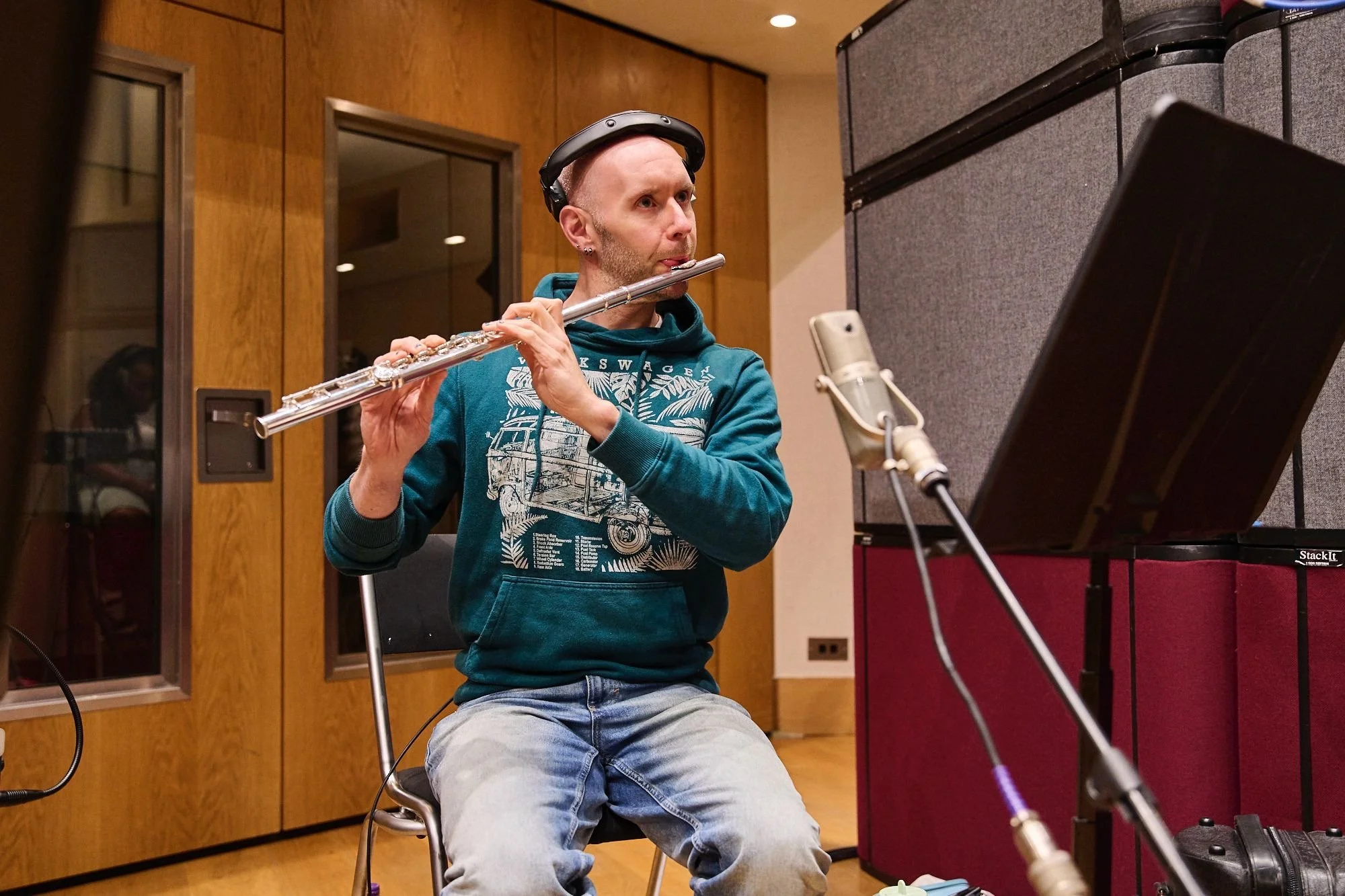 A man with a shaved head, wearing a teal hoodie and jeans, sitting on a chair in a recording studio while playing a flute. He has a pair of headphones resting on his head and appears focused. There is a professional microphone setup in front of him and a music stand to his right. The room has wood-paneled walls and acoustic panels.