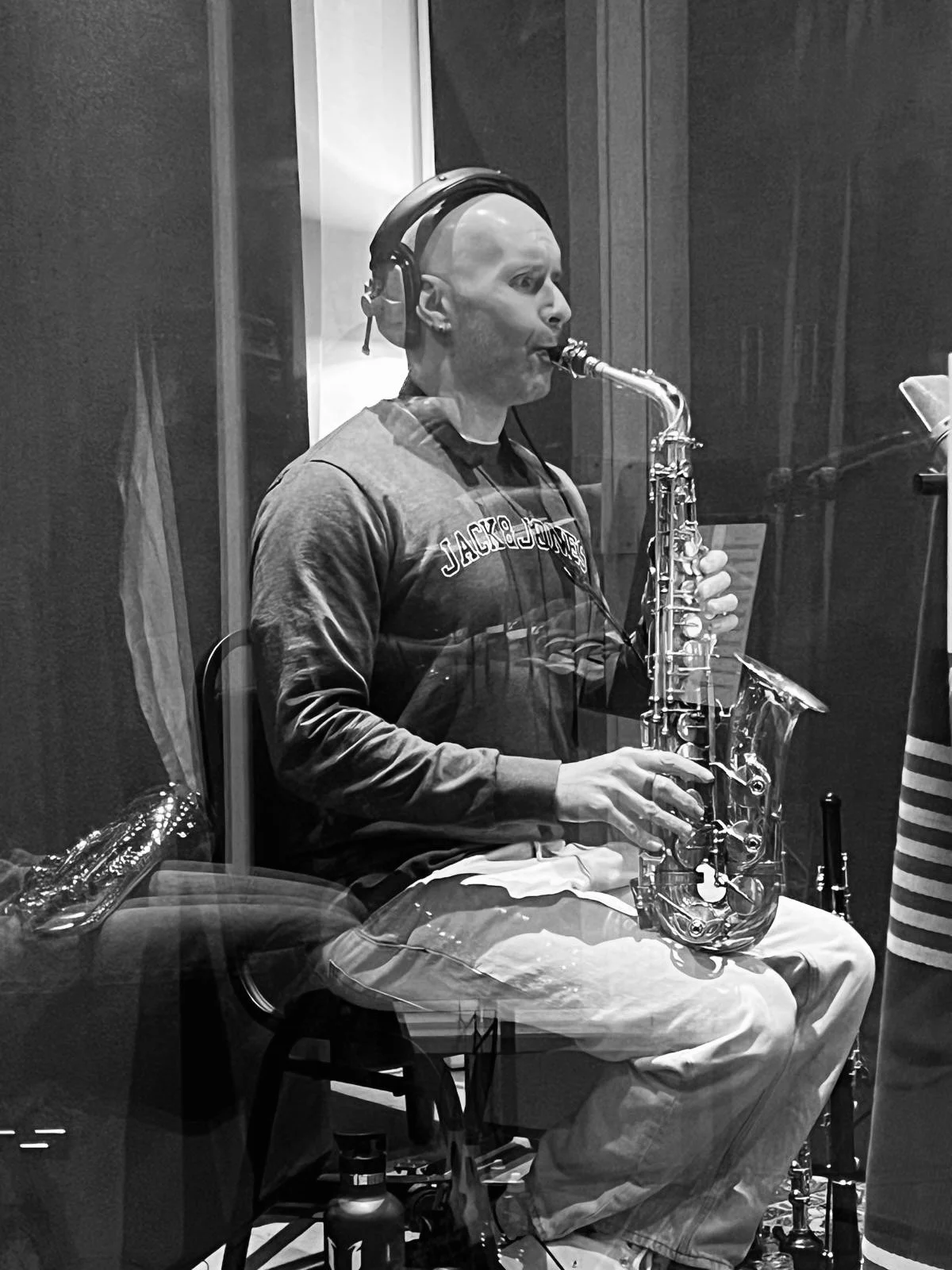 Man playing saxophone in recording studio, wearing headphones, sitting behind a glass window.
