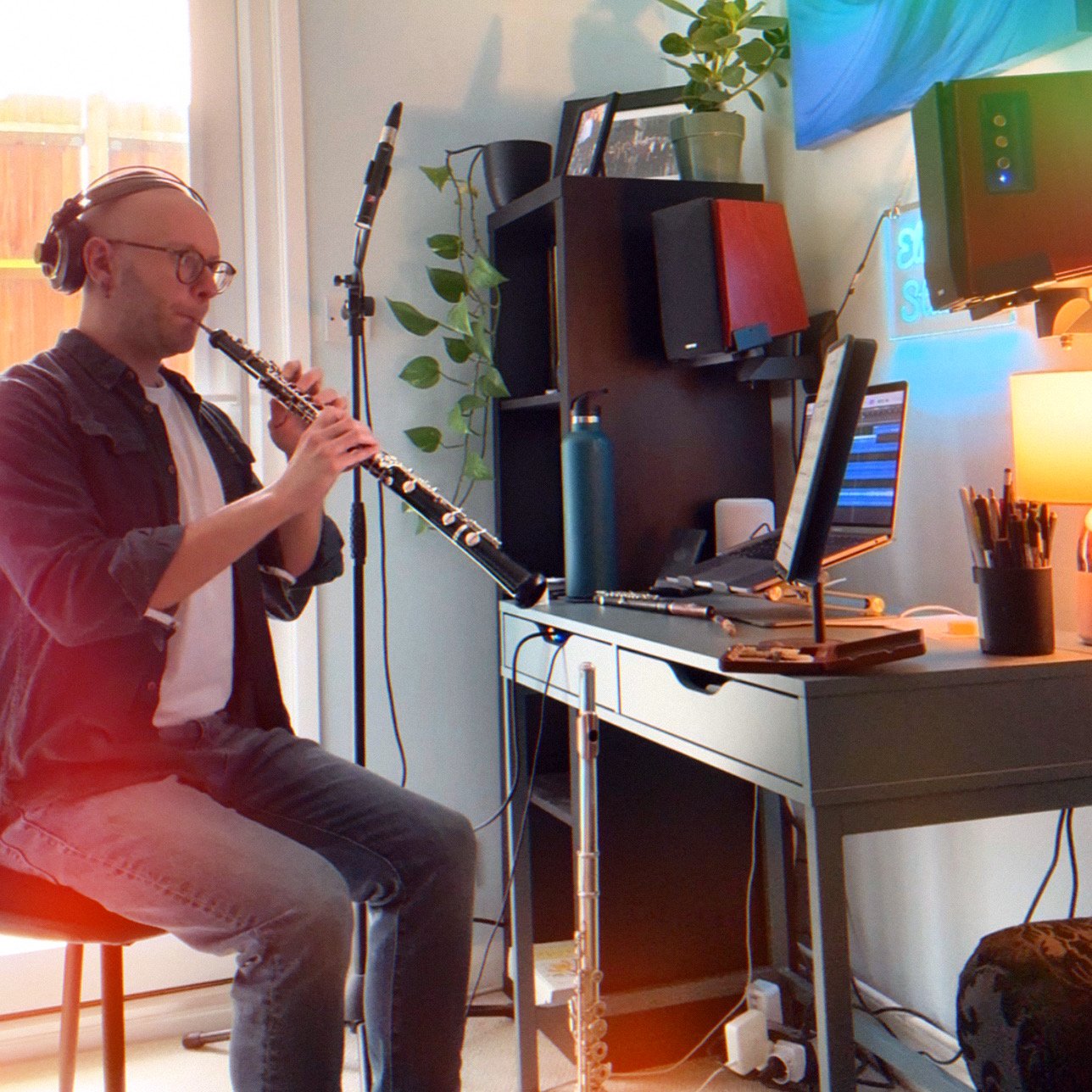 A man playing the oboe in a home studio with musical equipment, computer, and plants.