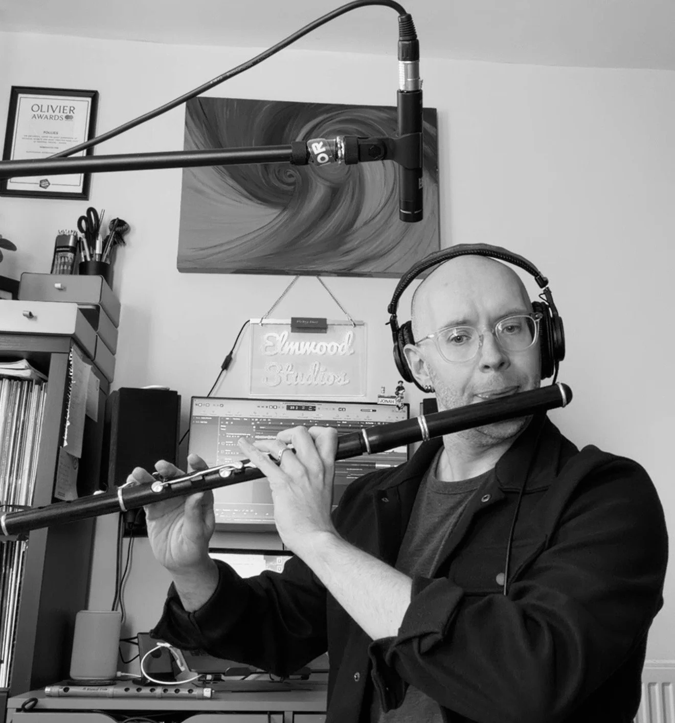 A man with glasses and a headset playing a flute in a home recording studio with a computer, a monitor, a neon sign, and various art and office supplies in the background.