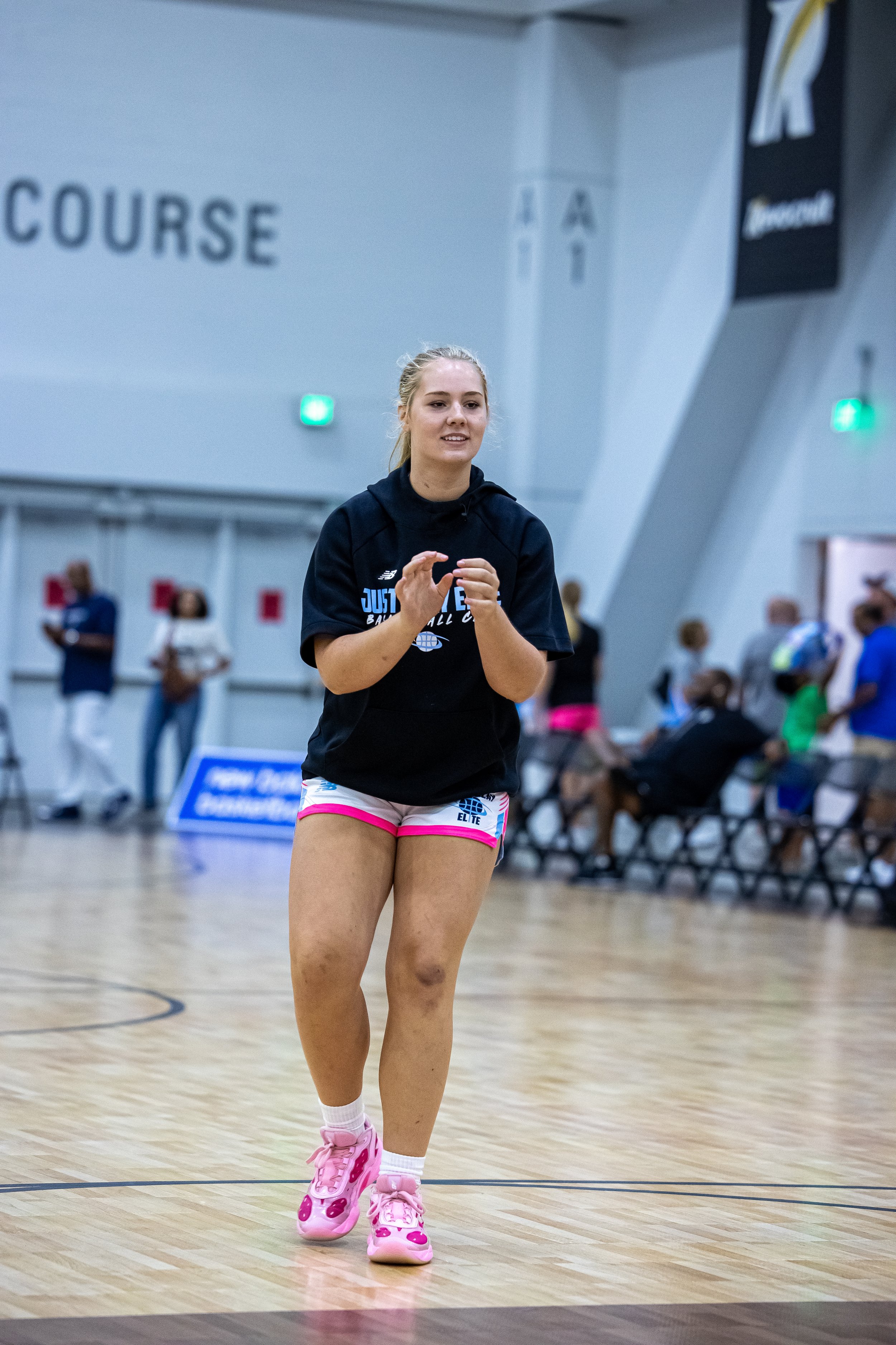 A female basketball player dressed in a white, blue, and black uniform with the phrase "JUST PLAY" and the number 1, standing on an indoor basketball court.