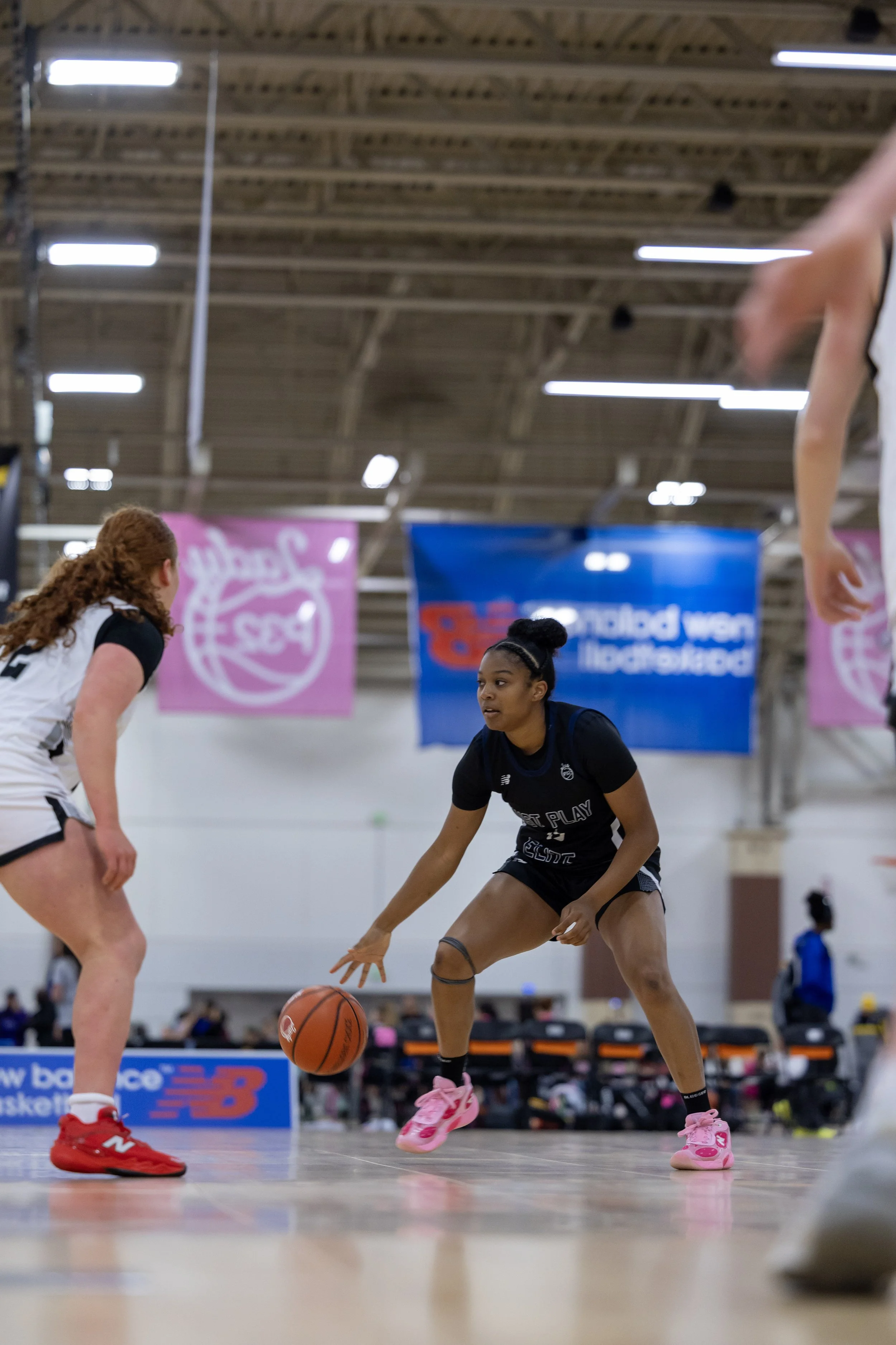 Four female basketball players contest for the ball during a game, with a player in a white jersey holding the ball surrounded by three players in black jerseys.