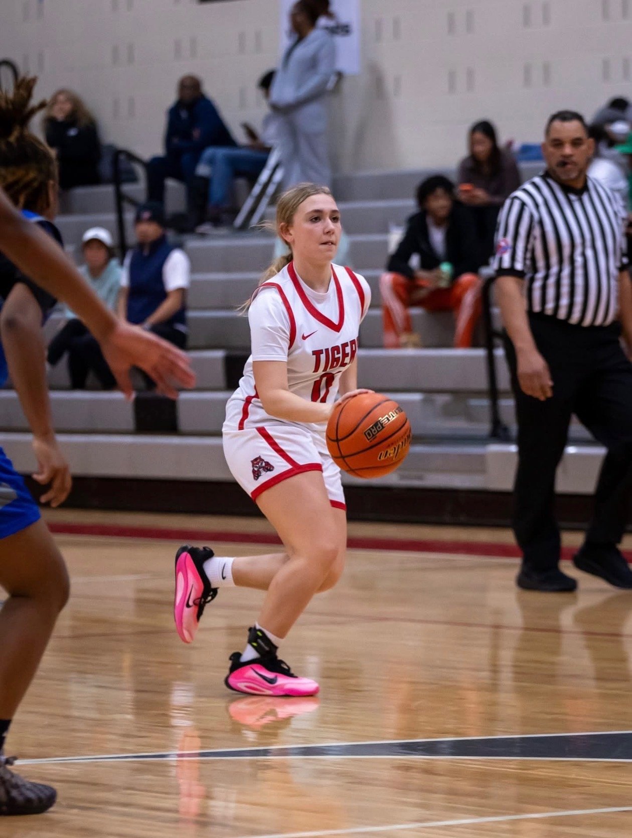 A female basketball player in a white jersey with red accents and the word 'TIGERS' dribbling a basketball down the court, with people seated on bleachers and a referee in the background.