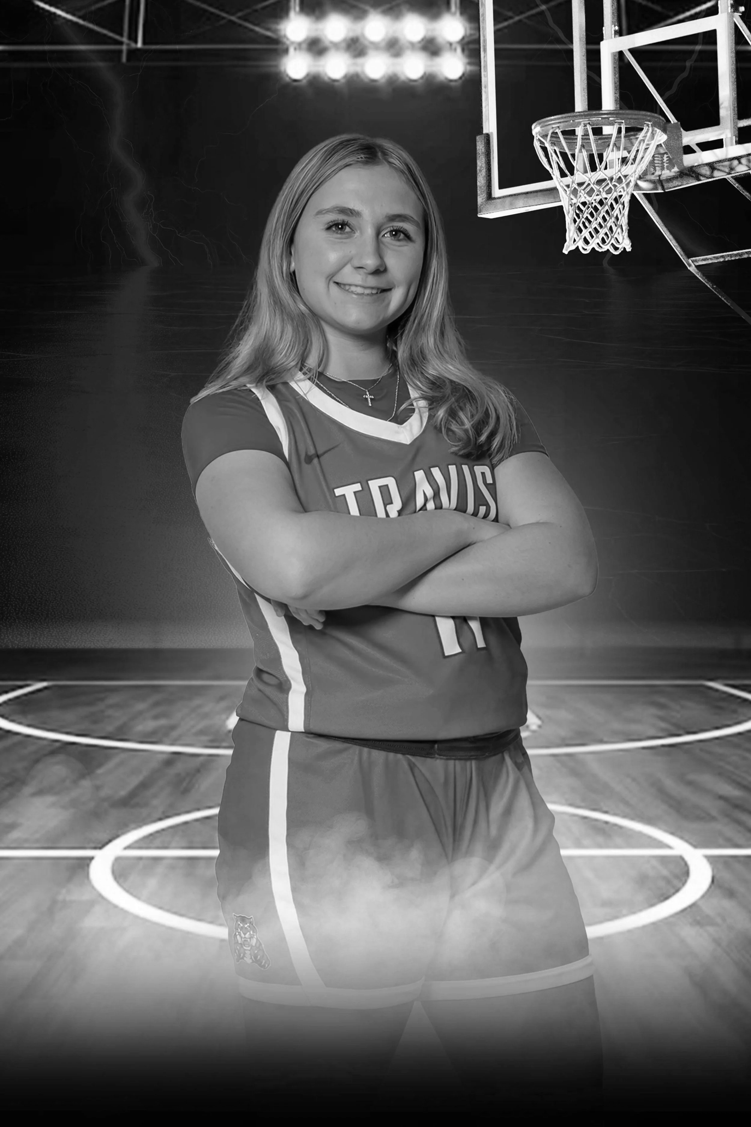 A young female basketball player in uniform standing with arms crossed on a basketball court, smiling, with a basketball hoop in the background.