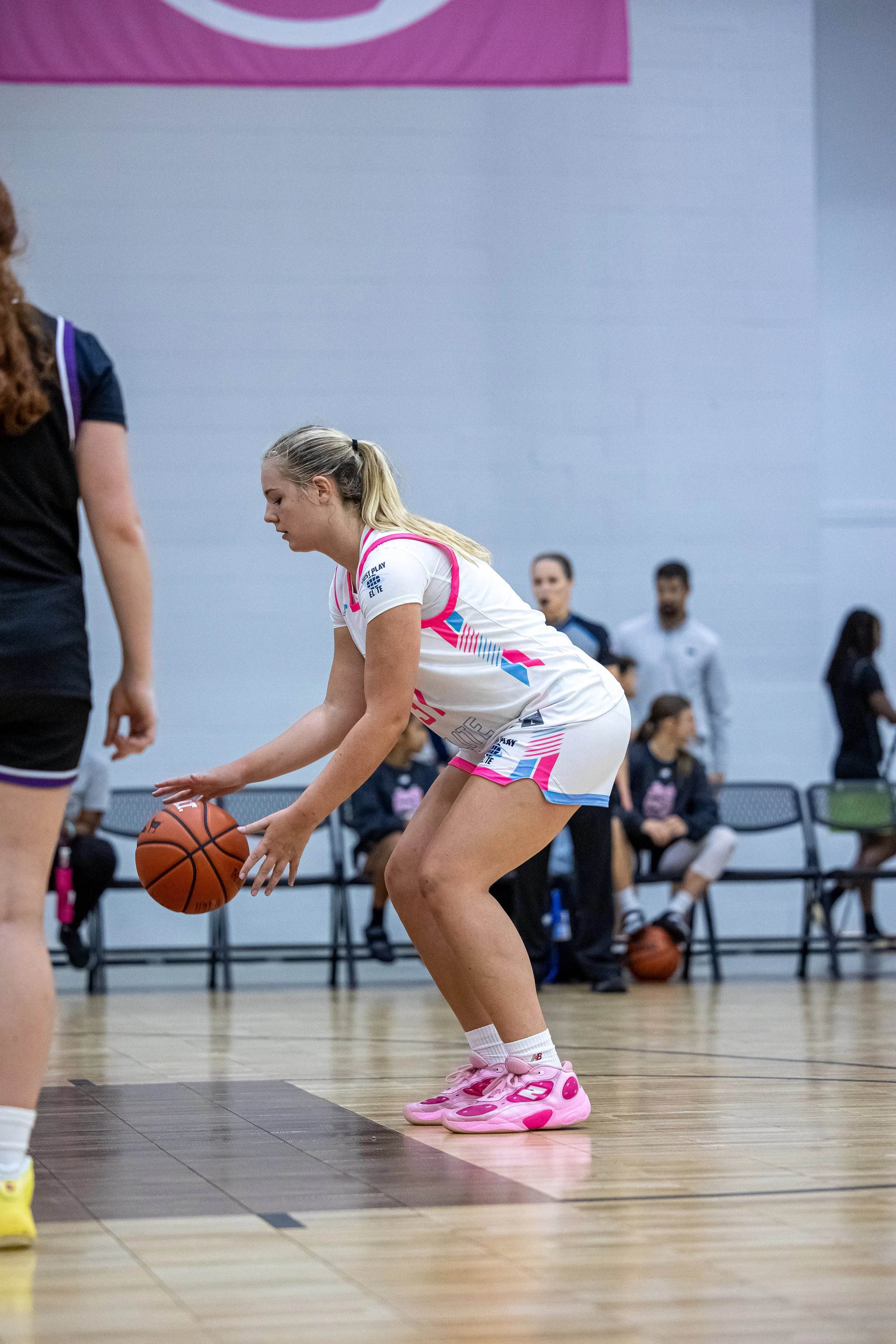 A female basketball player with long blonde hair in braids, wearing a blue and white uniform, prepares to jump for a rebound on an indoor basketball court.