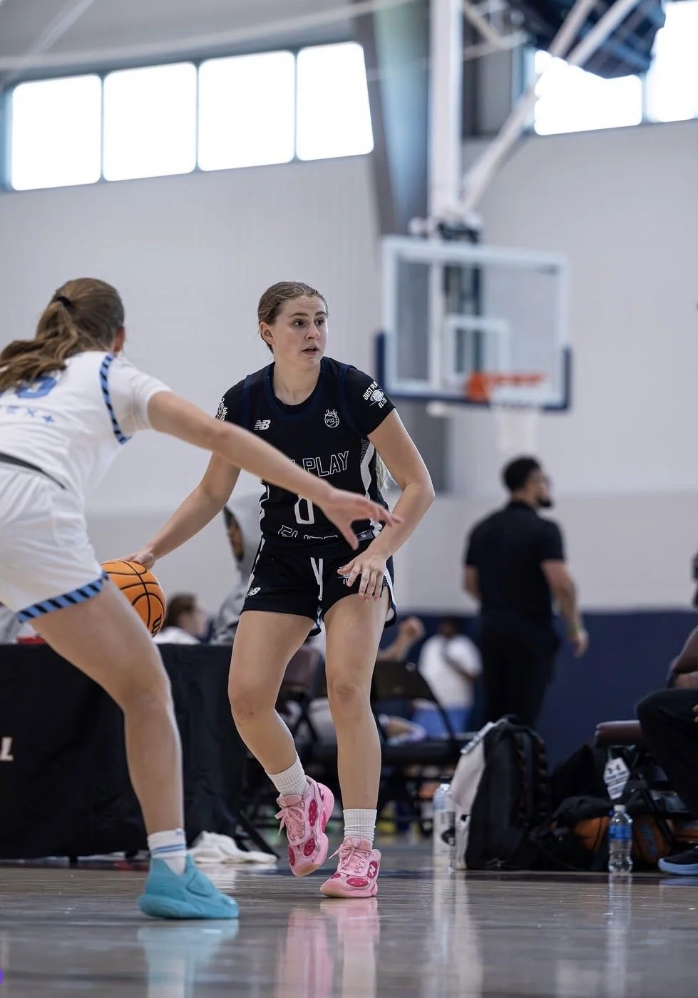 Two female basketball players competing for the ball during a game; one player wearing a white and blue uniform from 'Just Play' and the other in a dark blue and red uniform from 'Texas U'; the player in white is dribbling, while the player in blue is defending.