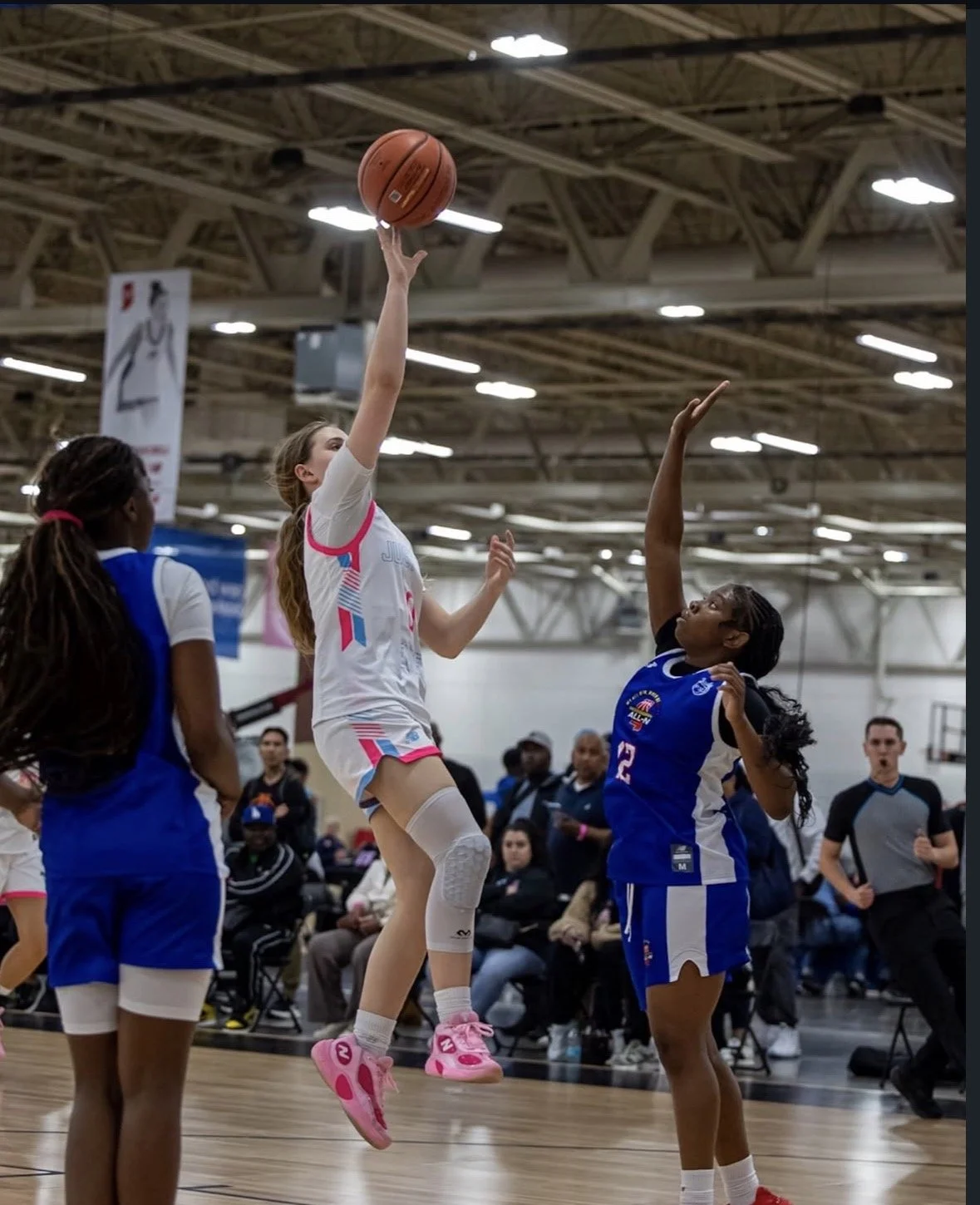 Three female basketball players on the court during a game, with one player dribbling the ball while two players attempt to defend her, in an indoor basketball arena.