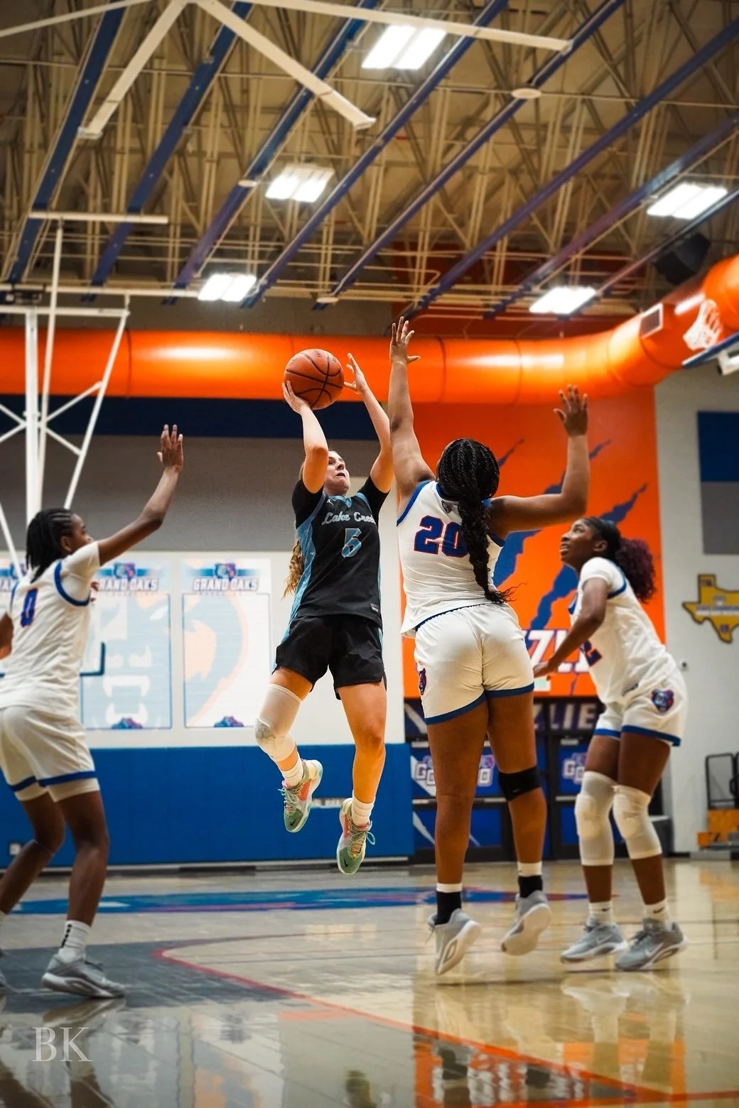Four female basketball players contest for the ball during a game, with a player in a white jersey holding the ball surrounded by three players in black jerseys.