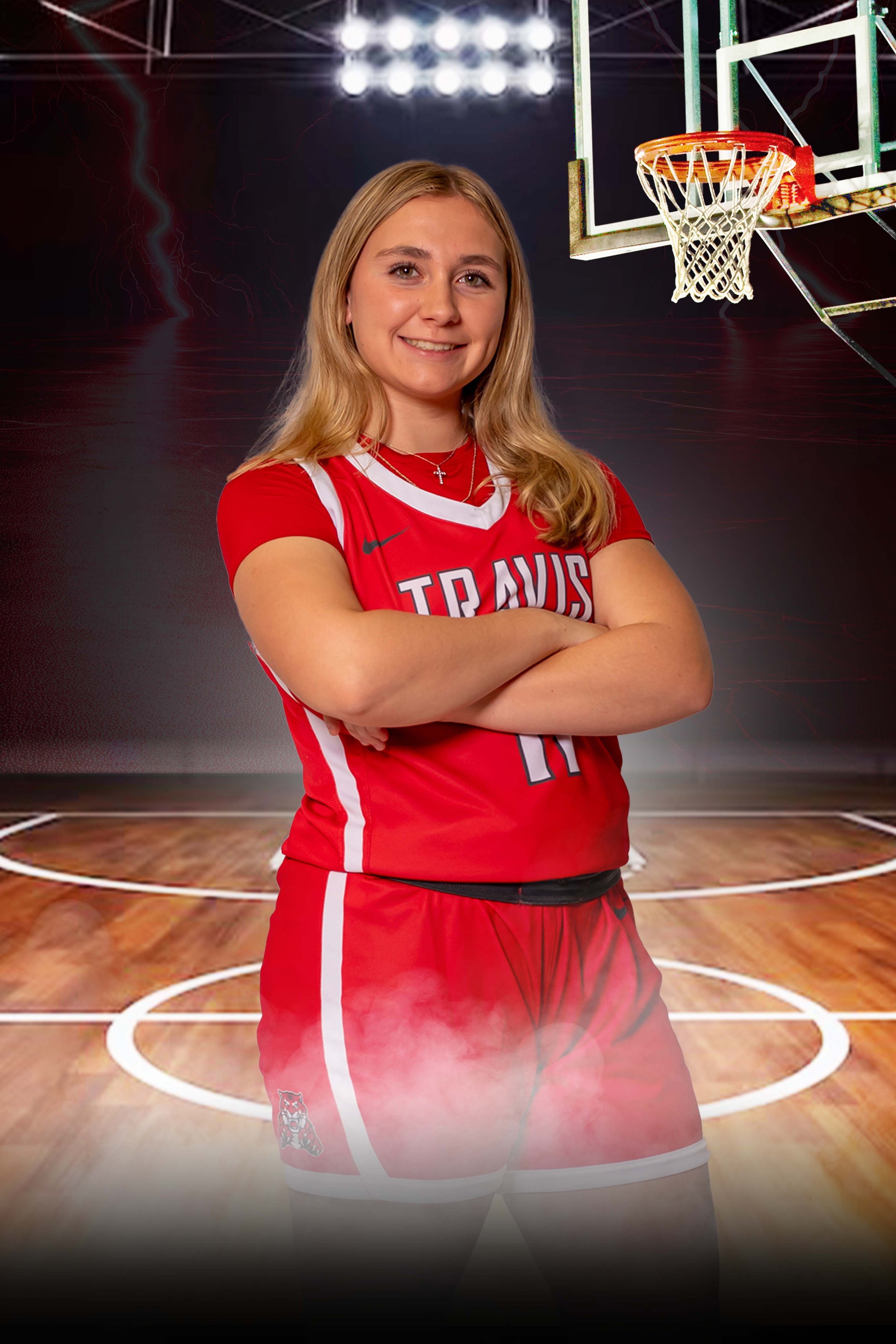 A young female basketball player in a red team uniform standing on a basketball court with arms crossed, smiling, with a basketball hoop in the background.
