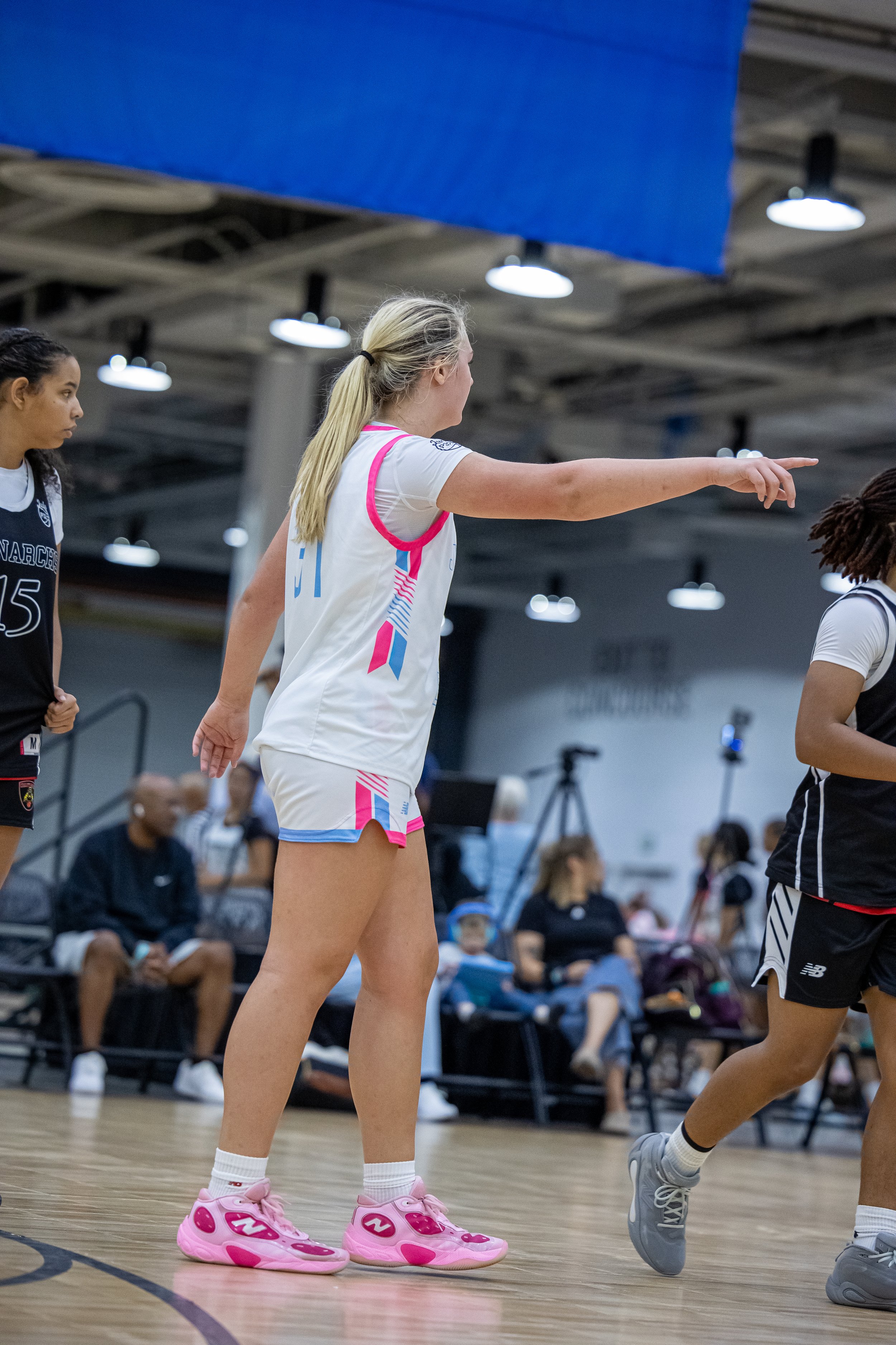 Three female basketball players on the court during a game, with one player dribbling the ball while two players attempt to defend her, in an indoor basketball arena.