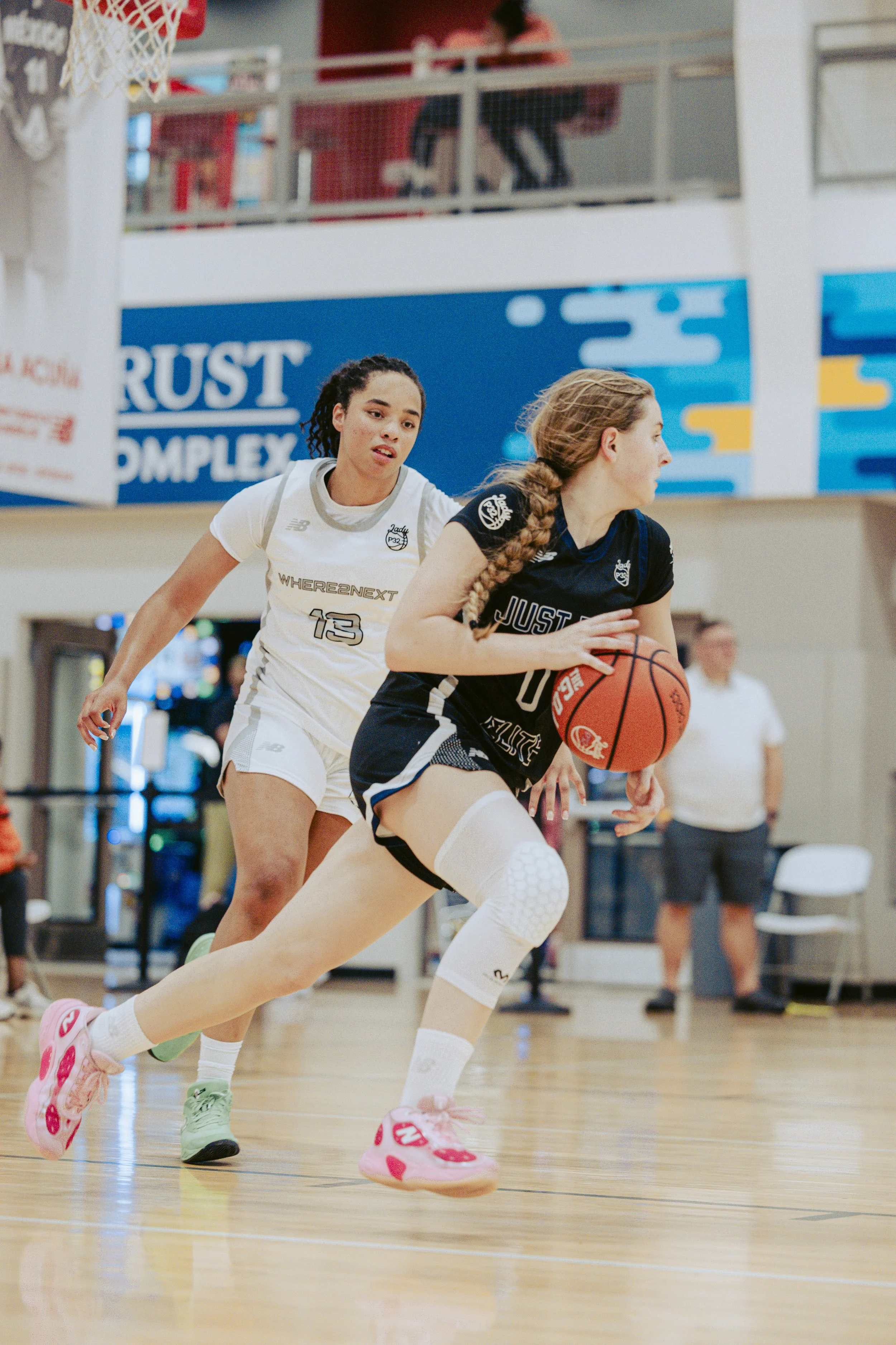 Two female basketball players compete on the court, one in a white and green uniform dribbling the ball, the other in a black and red uniform playing defense. The scene appears to be during a game in an indoor gymnasium.