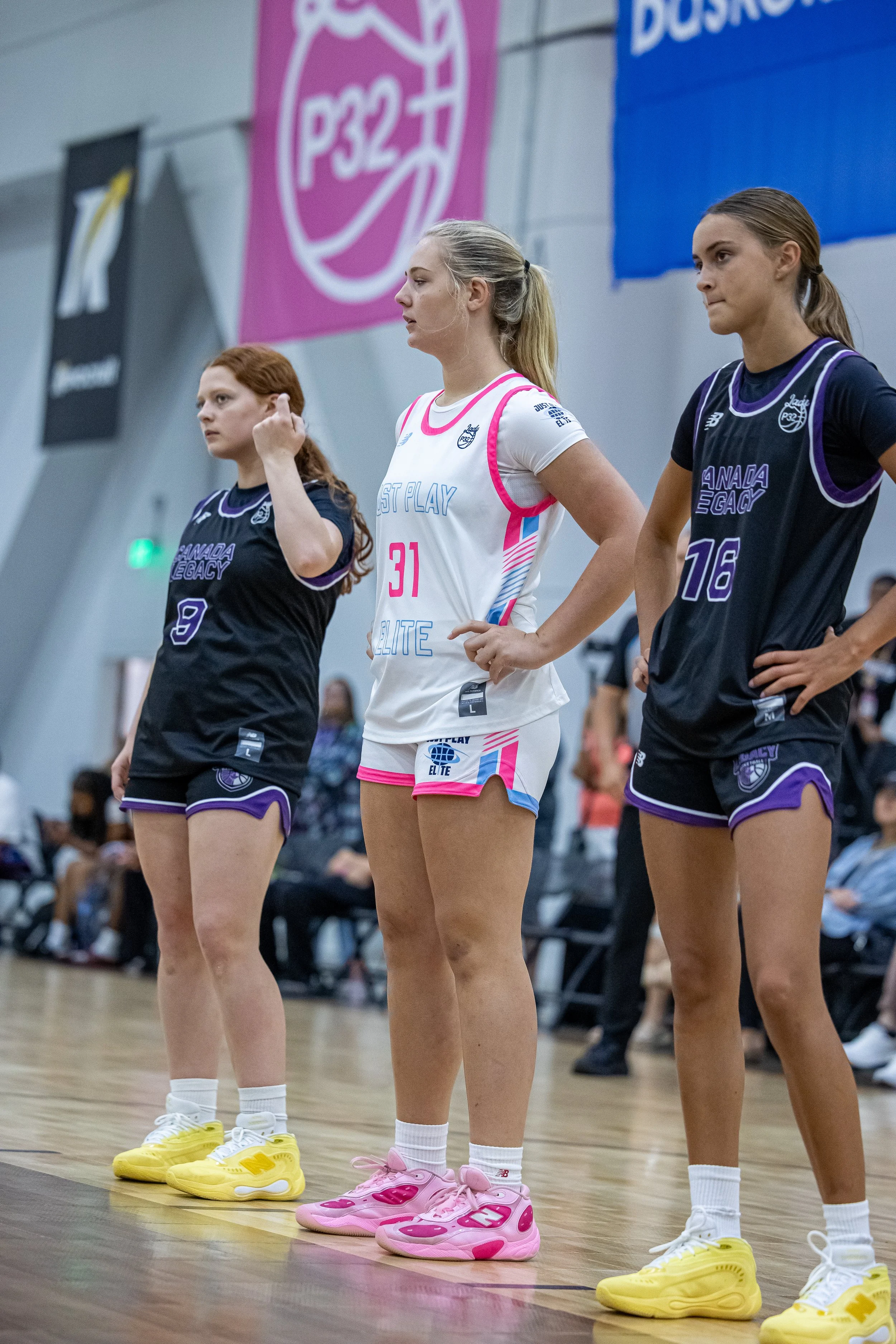 Four female basketball players contest for the ball during a game, with a player in a white jersey holding the ball surrounded by three players in black jerseys.