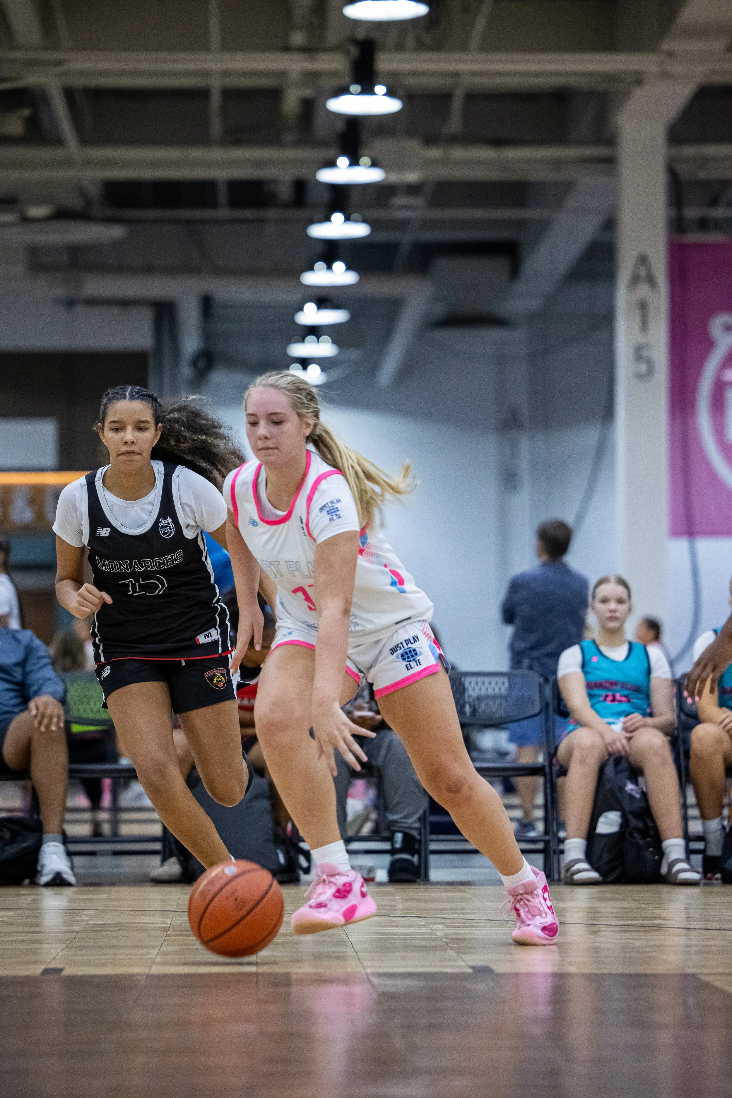 Two female basketball players competing for the ball during a game; one player wearing a white and blue uniform from 'Just Play' and the other in a dark blue and red uniform from 'Texas U'; the player in white is dribbling, while the player in blue is defending.