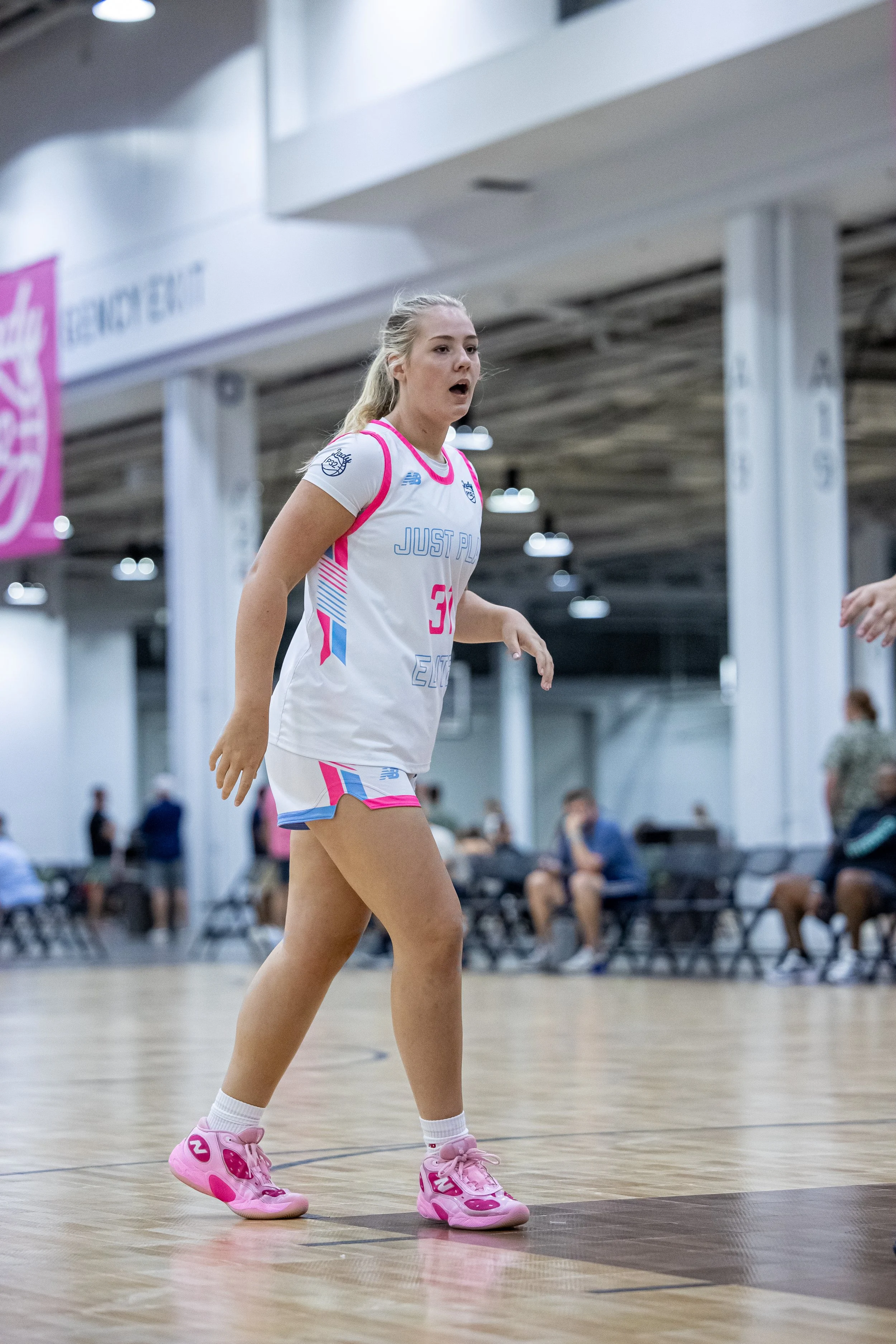 Two female basketball players compete on the court, one in a white and green uniform dribbling the ball, the other in a black and red uniform playing defense. The scene appears to be during a game in an indoor gymnasium.