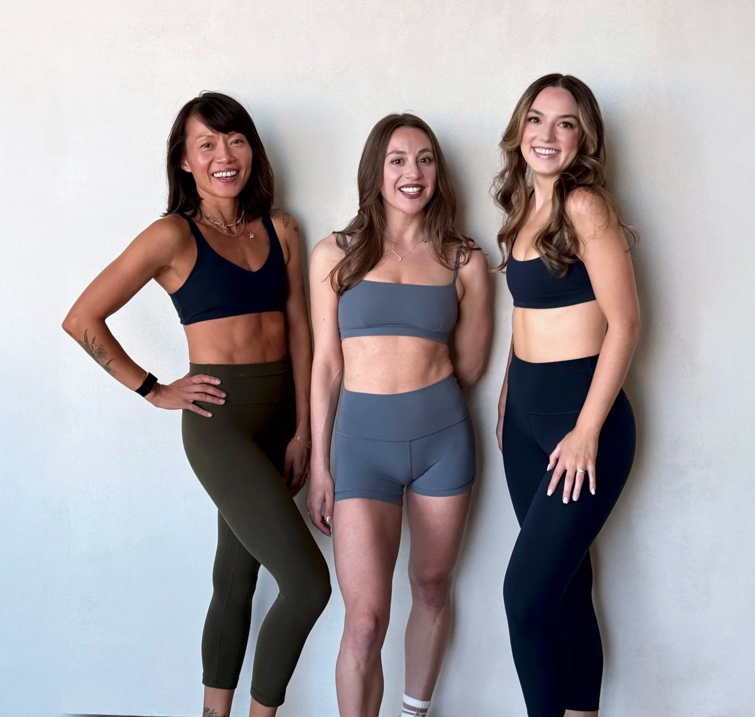 Three women smiling and standing in workout attire against a light-colored wall.