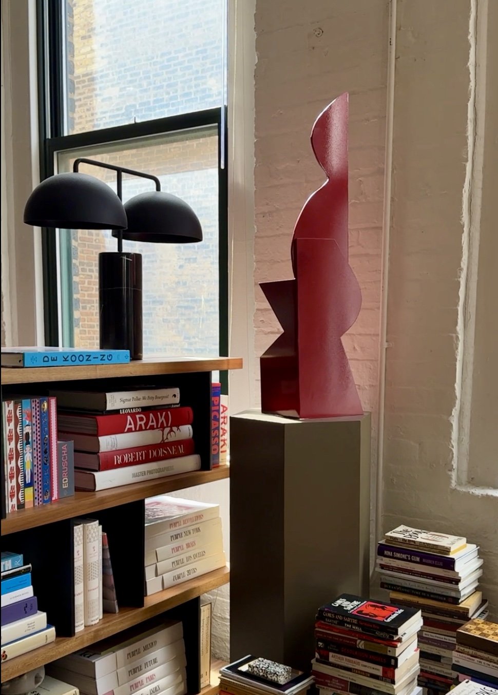 Interior scene with a modern black lamp on a bookshelf, a red abstract sculpture on a pedestal, and stacks of books, positioned in front of a large window next to a brick wall.