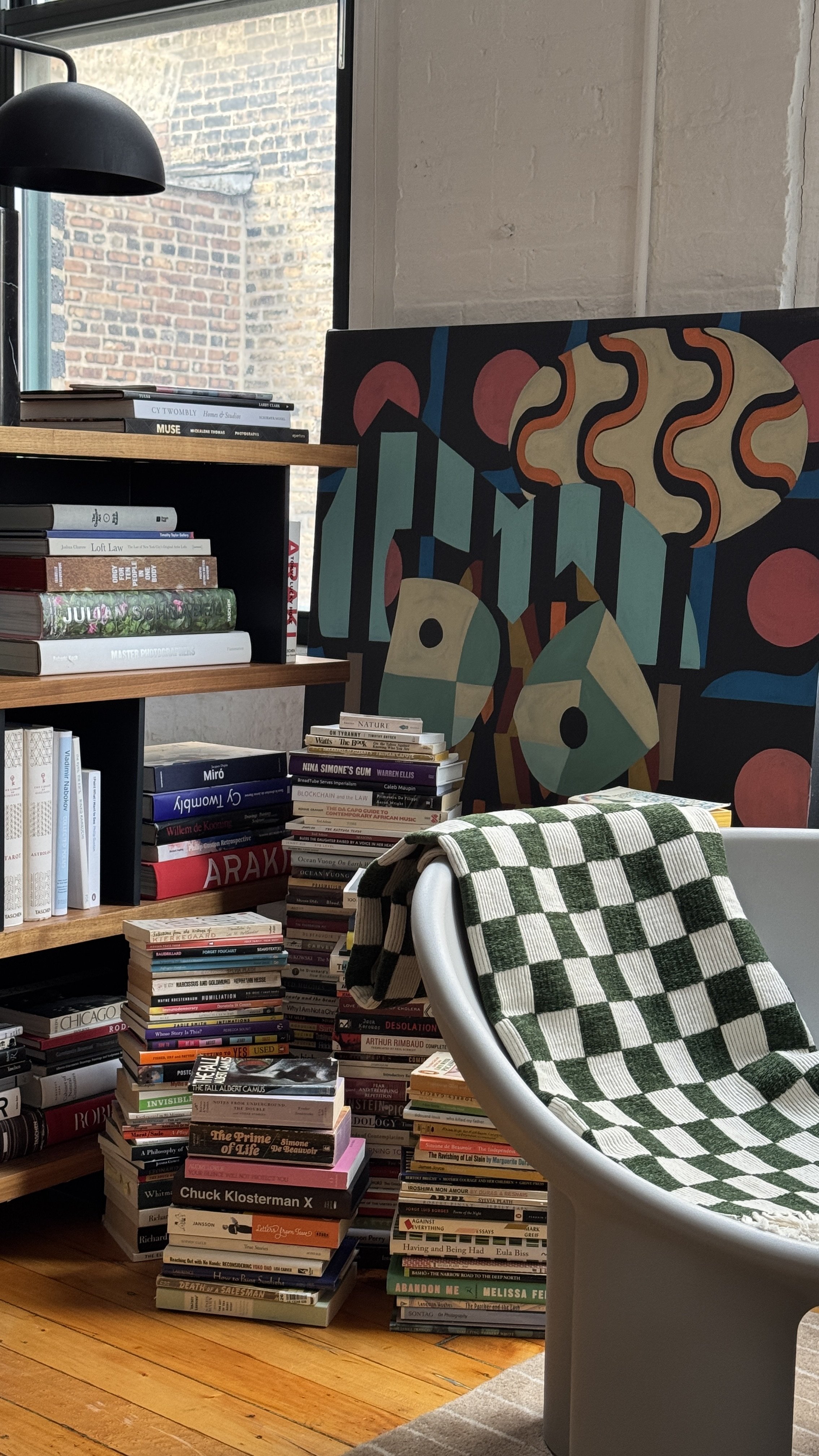 Cozy reading nook with a stack of books and a modern chair draped with a green checkered blanket. An abstract painting leans against a wall. Bookshelves are filled with various books and a window provides natural light.