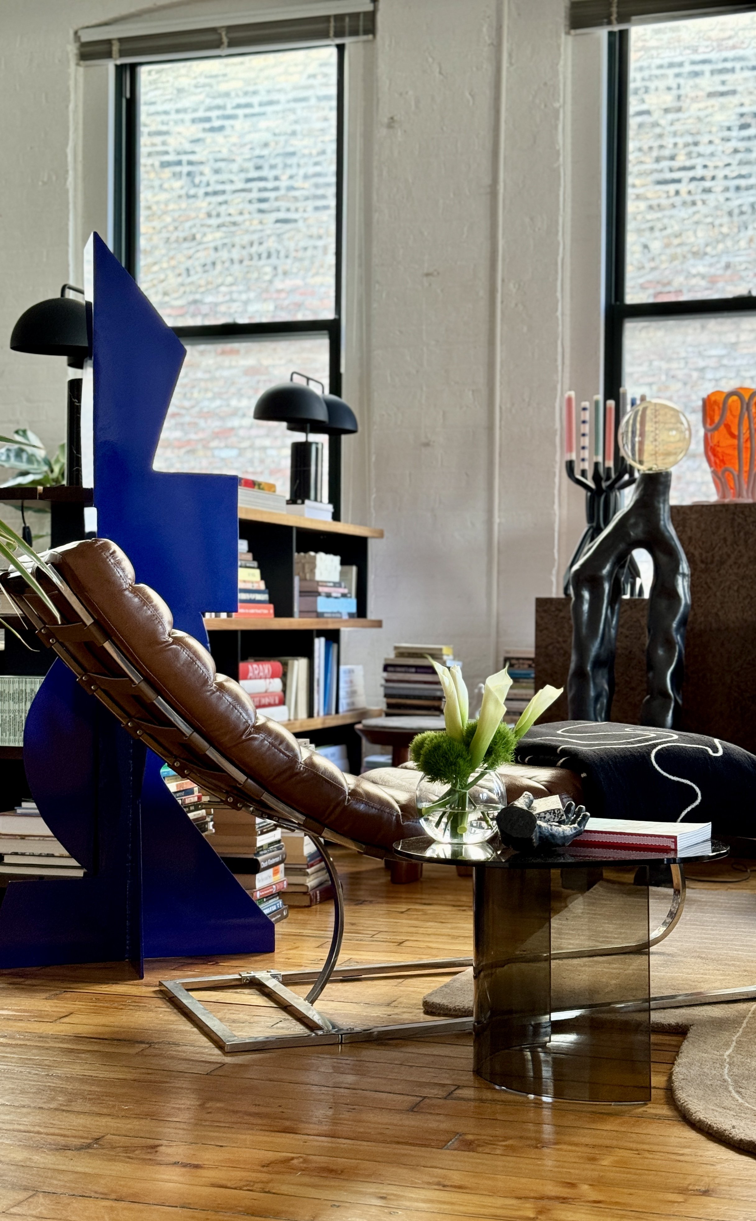 Modern living room with a brown leather chair, bookshelf with books, abstract art sculptures, large windows, and a glass coffee table with a vase of flowers.