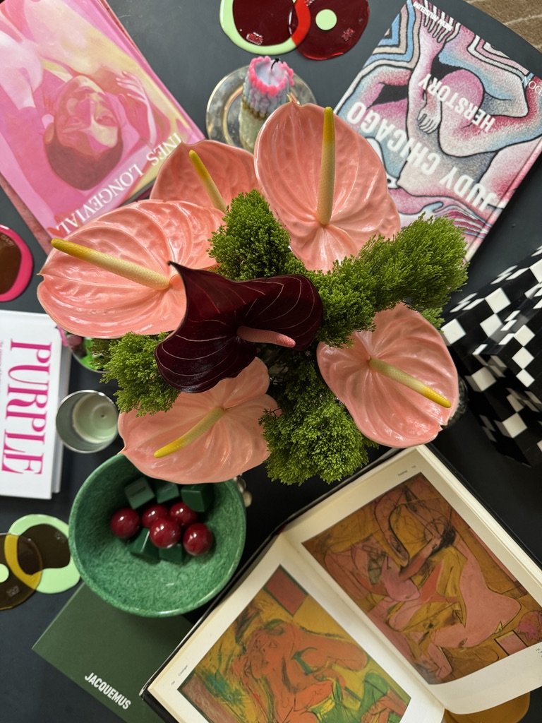 Art books and magazines on a table with pink anthurium flowers and decorative items.