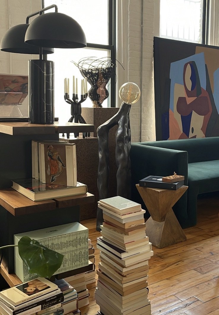 A stylish indoor space with a modern black lamp, sculpture, abstract art, an emerald couch, books, and a small wooden table.