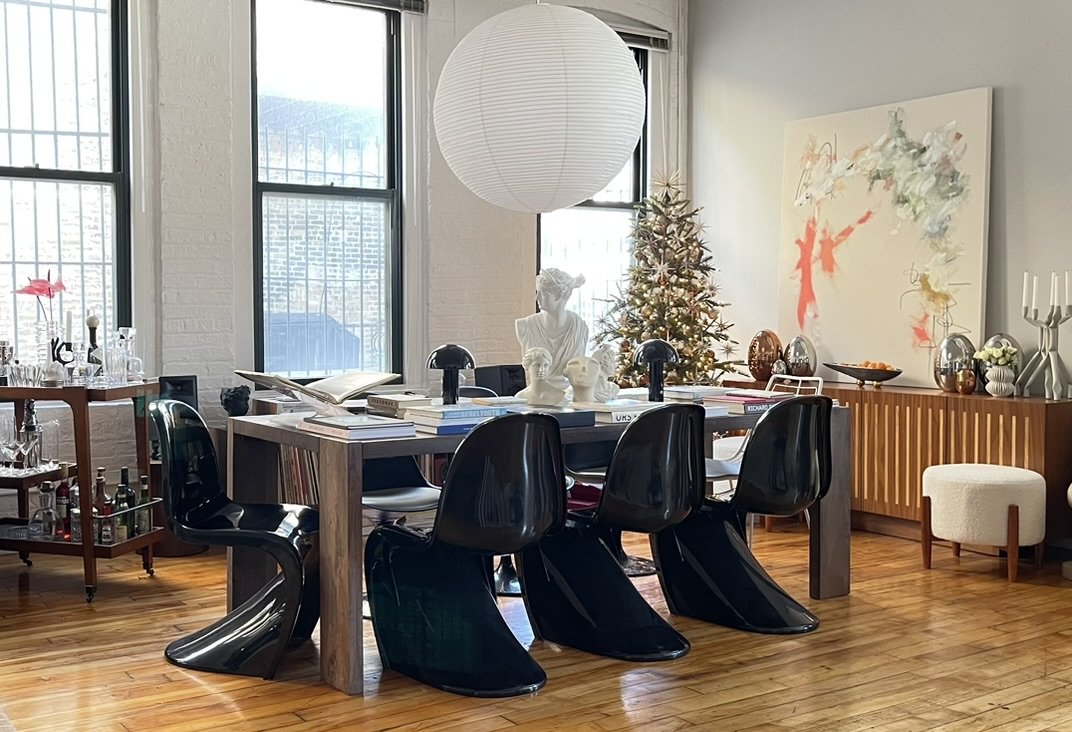 Modern dining room with black chairs, wooden table, books, art busts, and a paper lantern. A Christmas tree and abstract art painting are in the background. Bar cart with glassware is on the left.