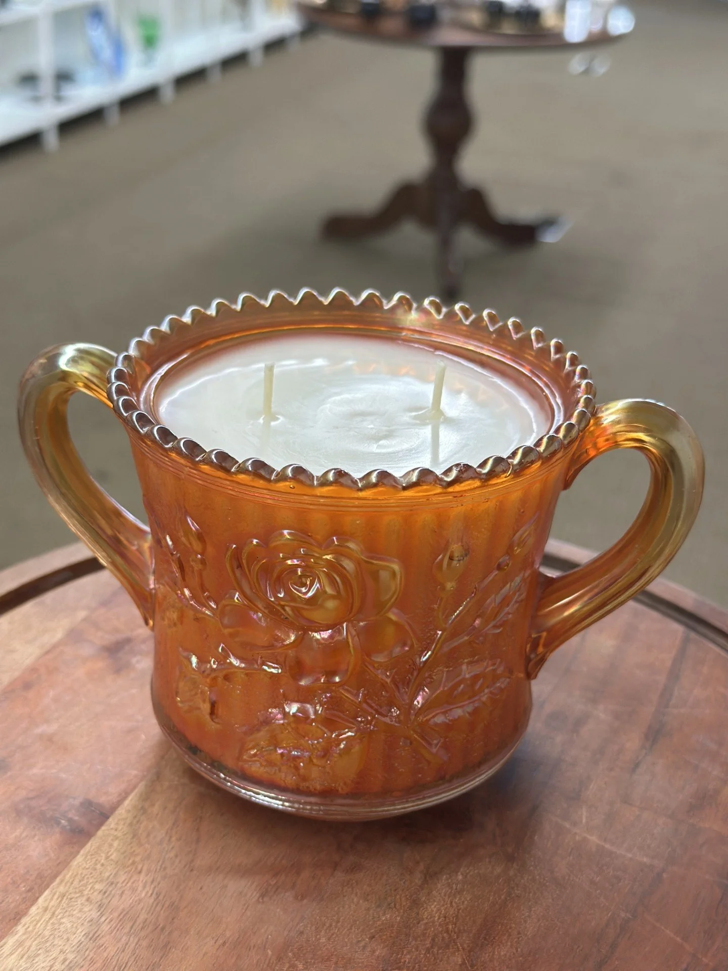 Amber-colored glass candle holder with hand poured candle inside, placed on a wooden surface, with a blurry background of a store or room.
