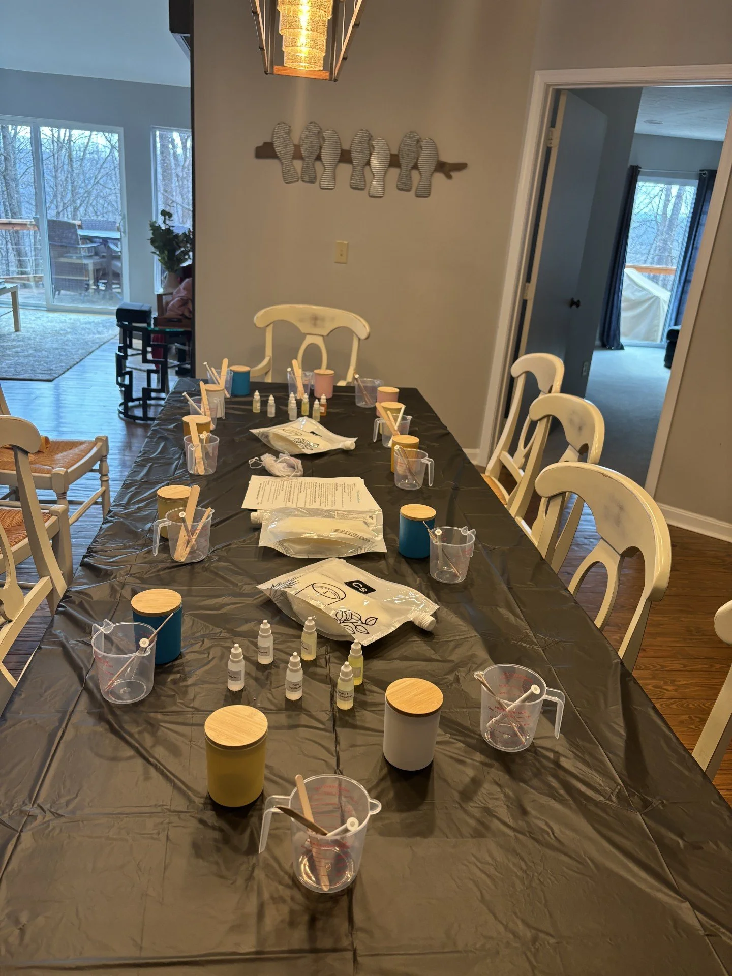 Candle-making supplies arranged on a dining table covered with a black tablecloth in a home setting with chairs around it.