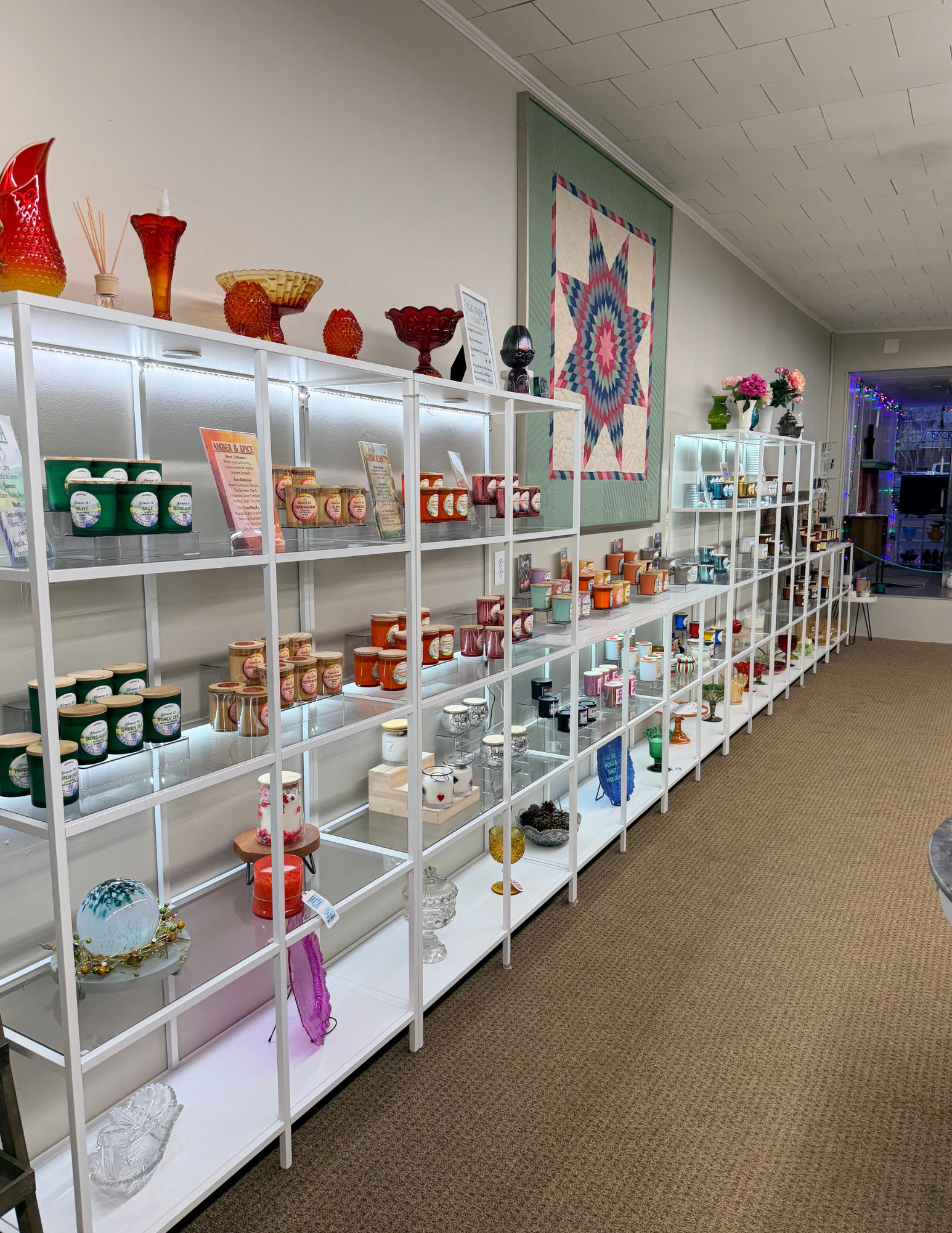 Display shelves with assorted candles and glassware in a store, with a quilted wall hanging and floral decorations in the background.