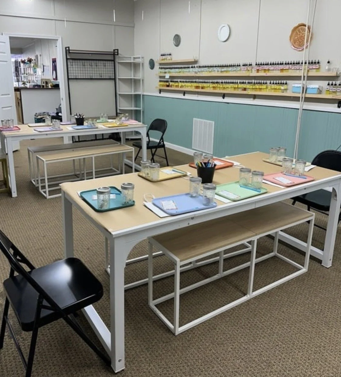 A classroom-style setup featuring tables with pastel-colored folders, glass jars, and writing utensils. Shelves lined with bottles are visible on the teal and beige walls. Foldable chairs and benches provide seating.
