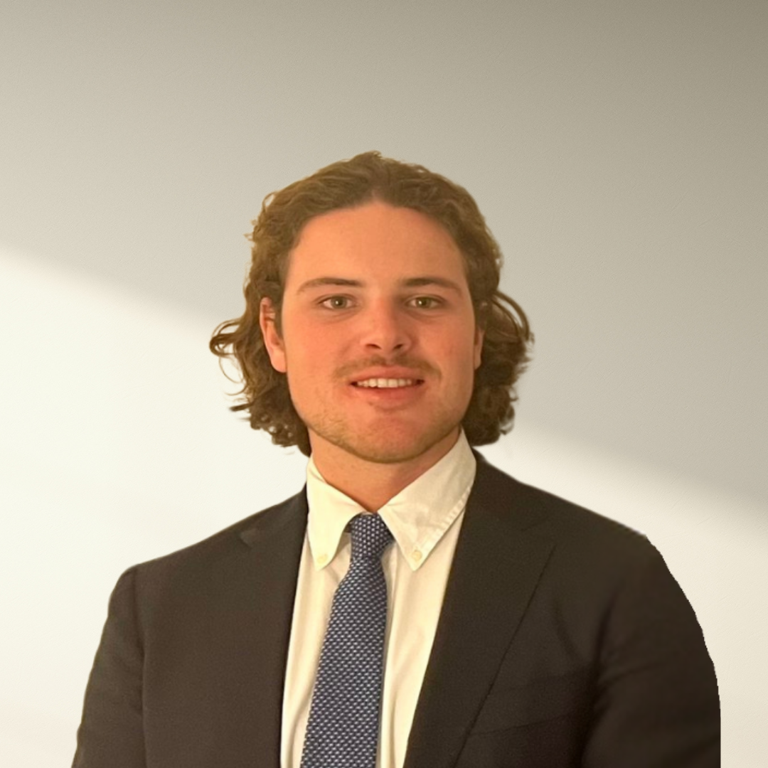 A young man with wavy brown hair dressed in a black suit, white shirt, and patterned tie smiling at the camera against a plain background with gradient lighting.