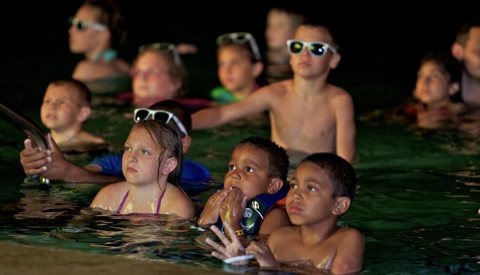 Children swimming underwater with goggles, some holding onto the pool's edge, in a dimly lit indoor pool.