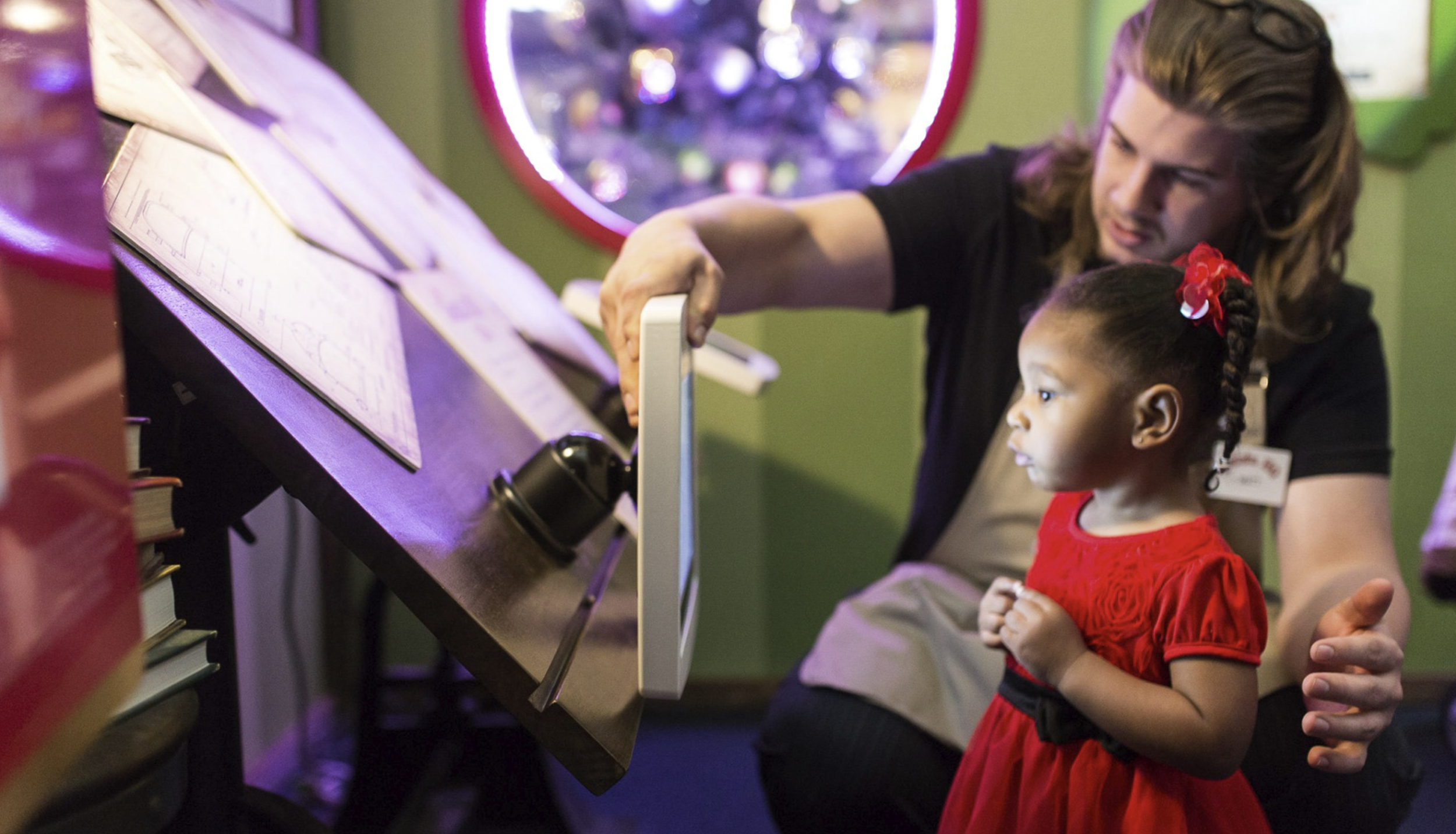 A young girl in a red dress with a red hair bow standing at a museum exhibit with an adult woman, pointing at an informational display and looking attentive.
